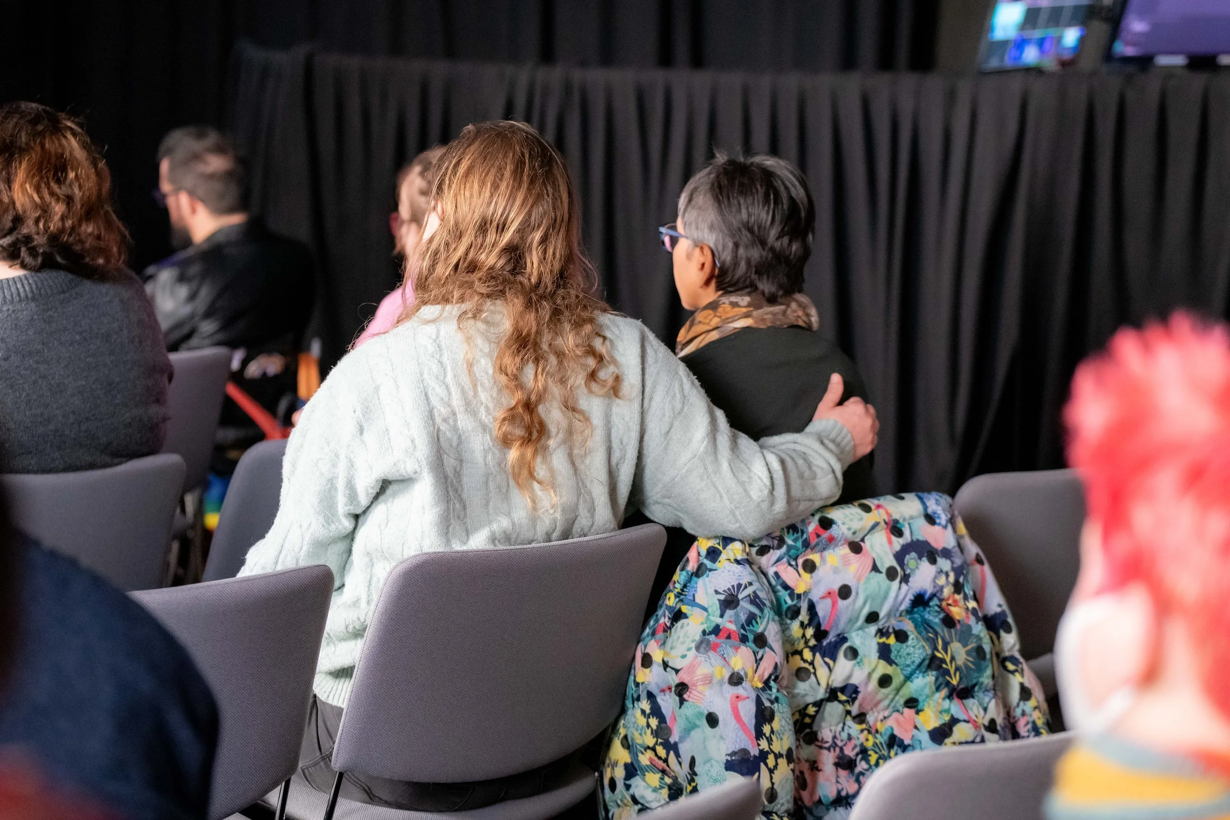 An audience member in a grey jumper hugs another human wearing a scarf.