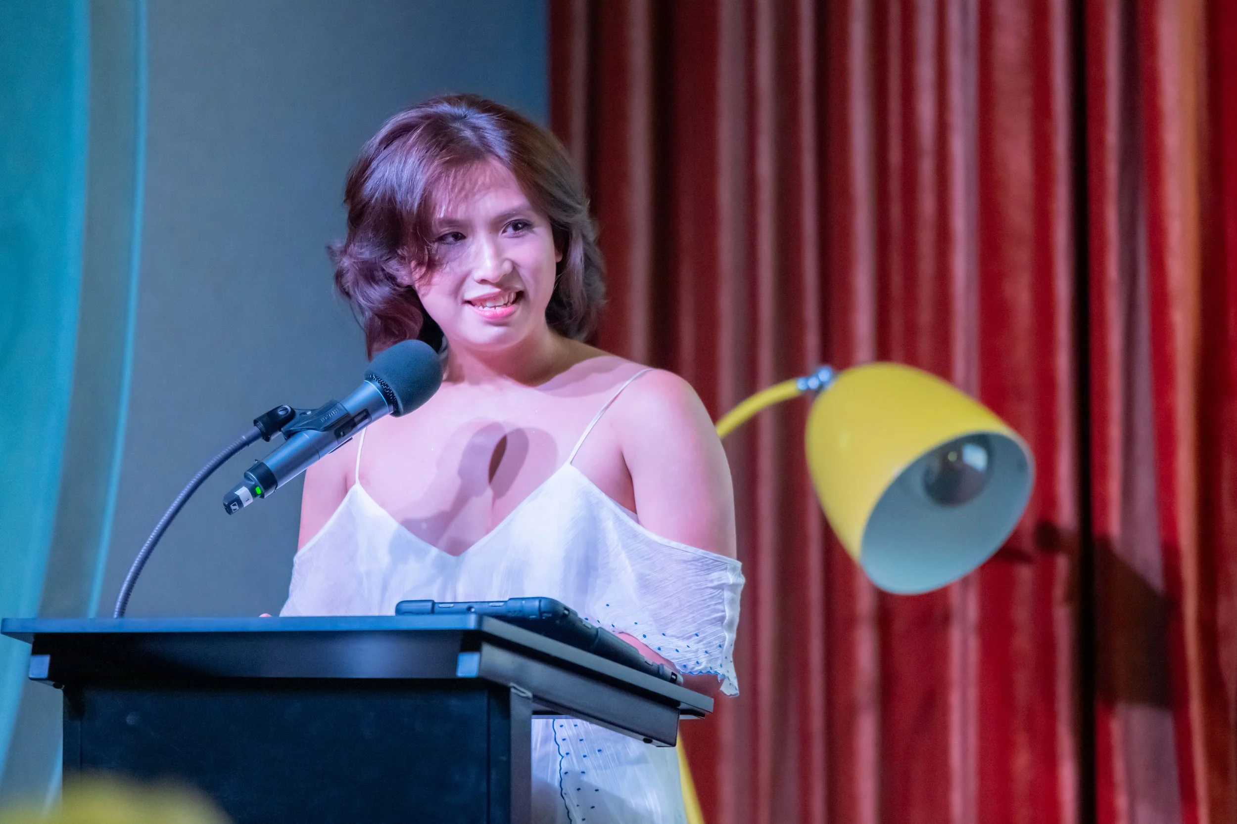 A human in a white dress stands at a lecturn and smiles.