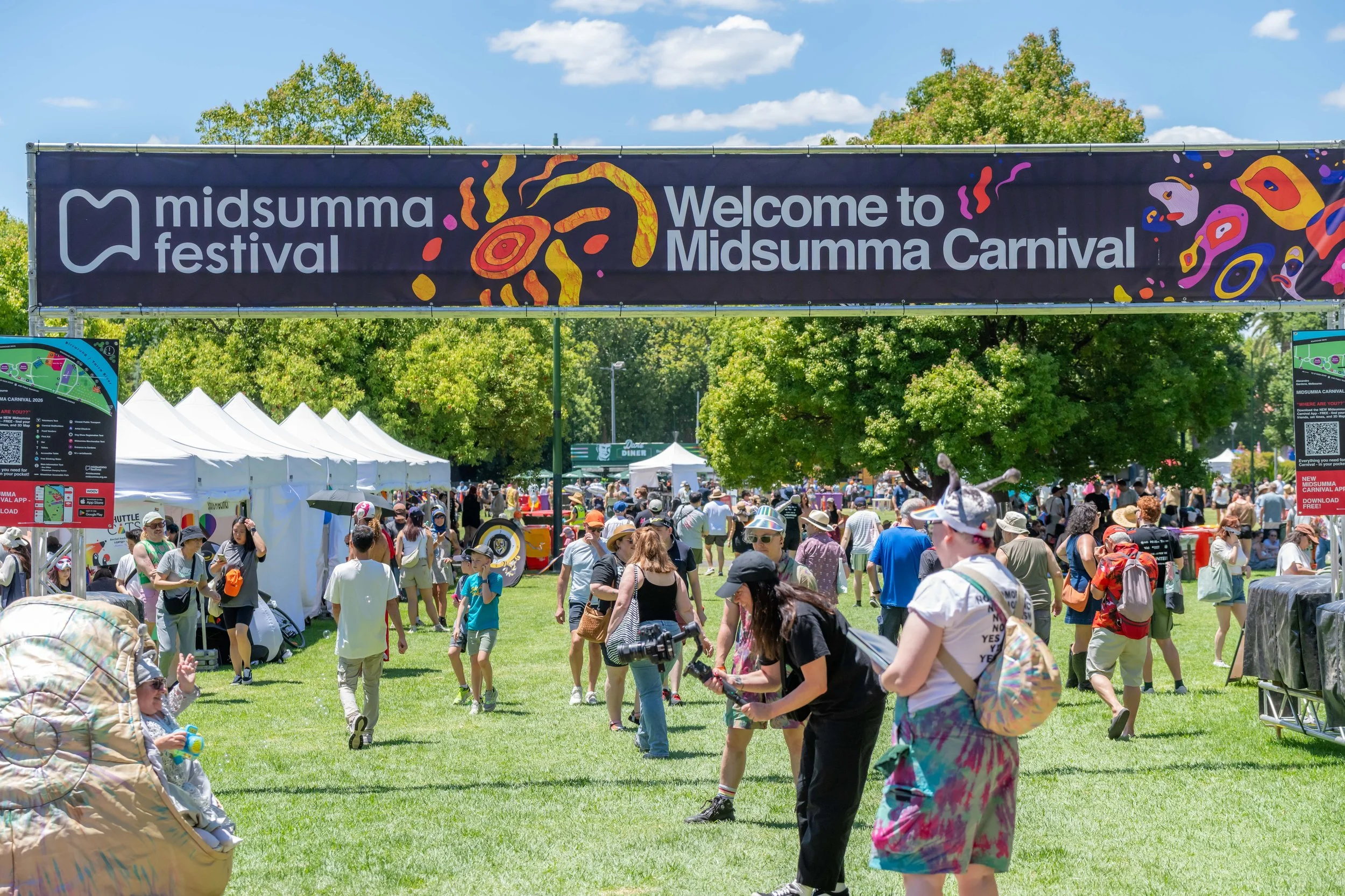 Many people mill about underneath a Welcome to Midsumma Carnival banner. The snail human is being photographed.