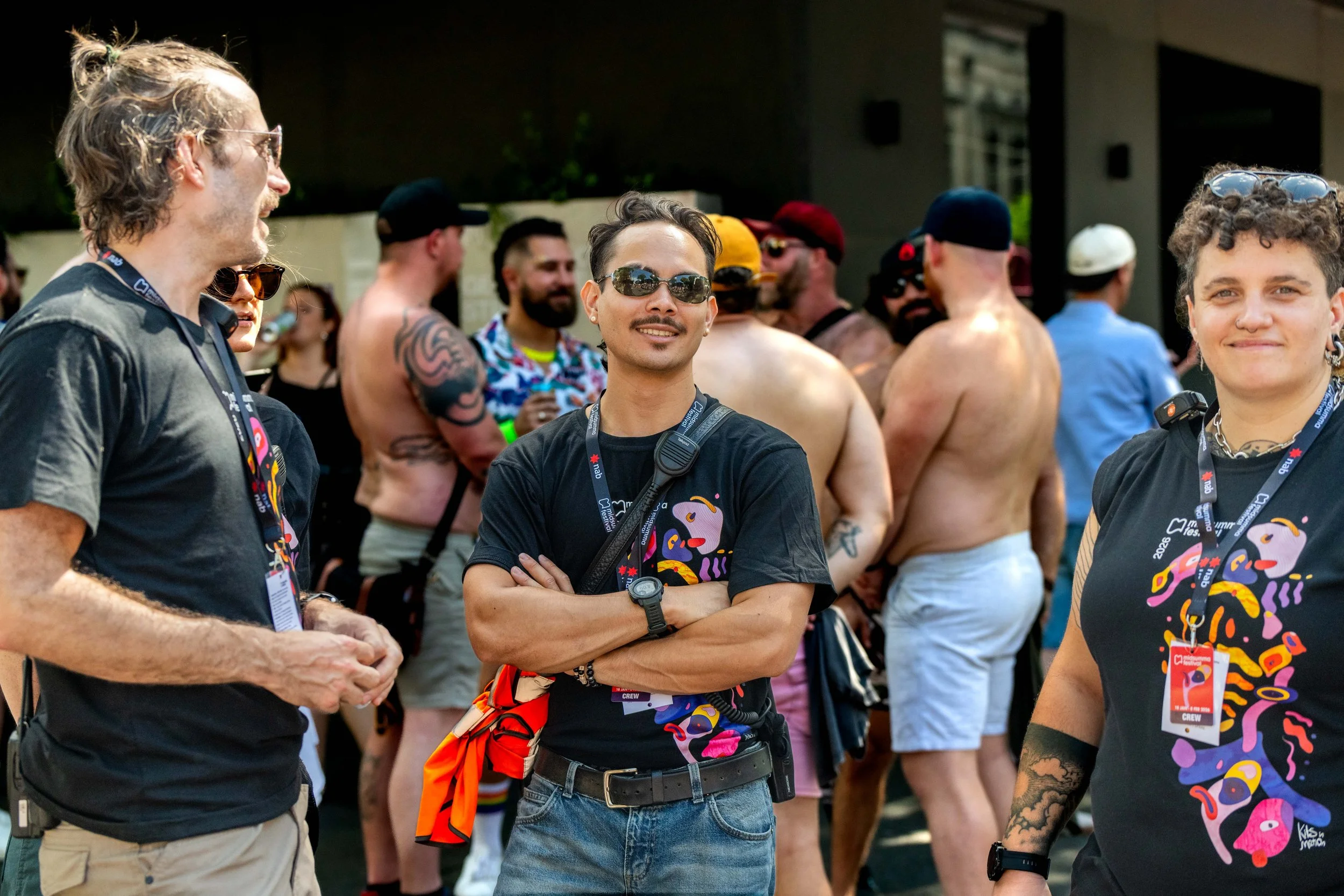 Three Midsumma Festival volunteers stand amongst a group of people in the street and chat.