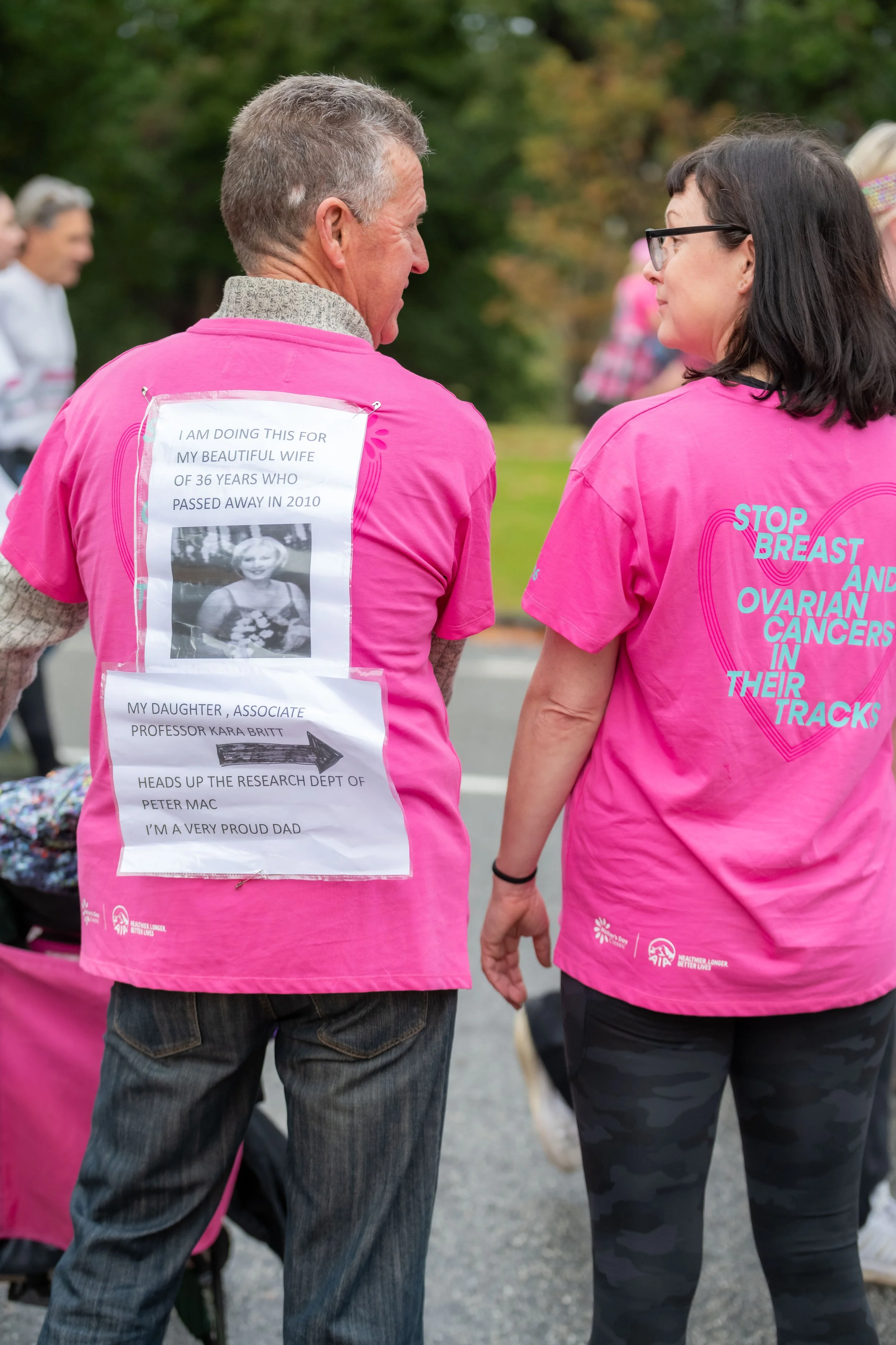 Two humans stand facing away from the camera. The racegoer on the left has a memorial message pinned to their back. The other racegoer has "Stop breast and ovarian cancer in their tracks" written on the back of their pink tee.
