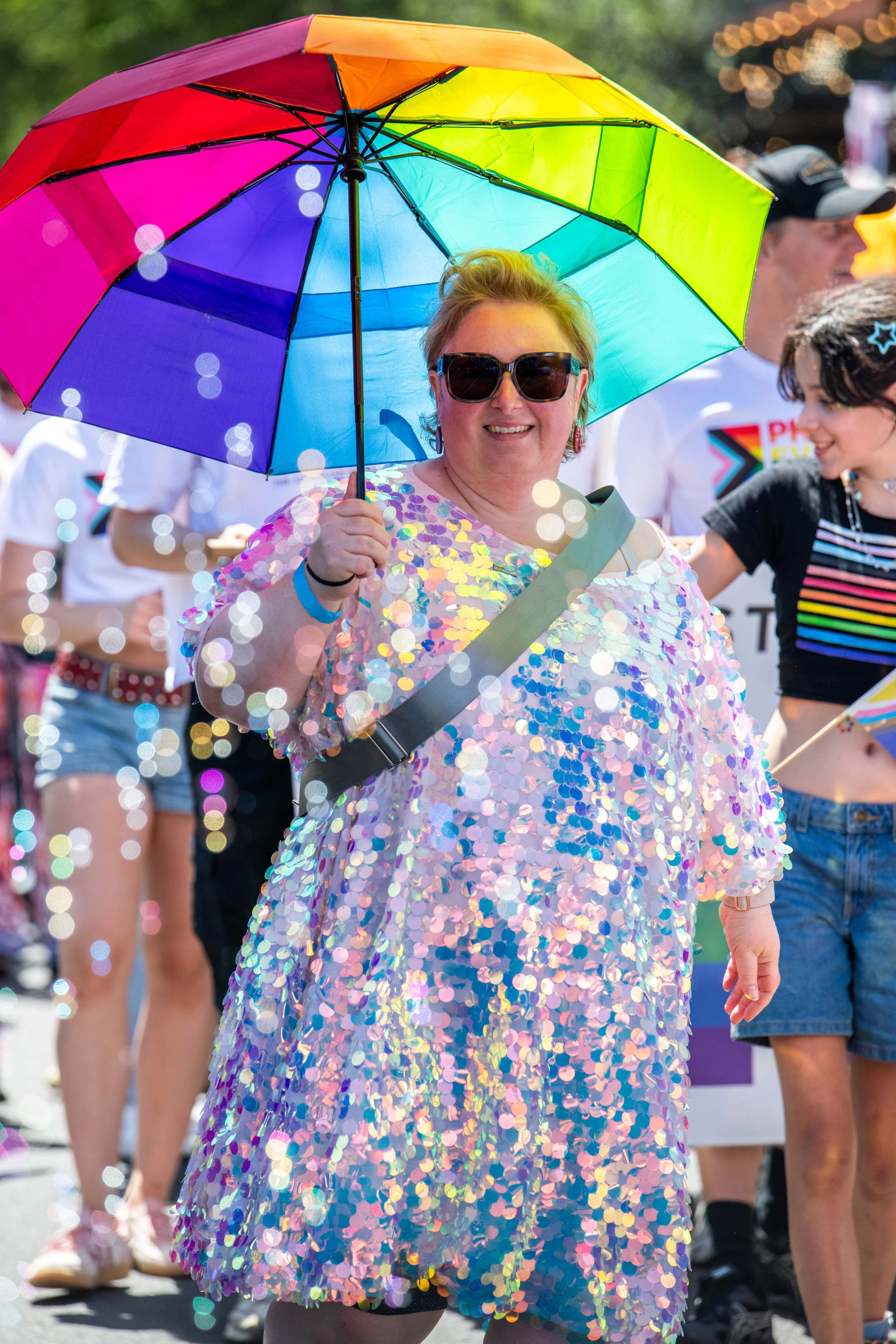 A human wearing a silver glitter dress and holds a rainbow umbrella.