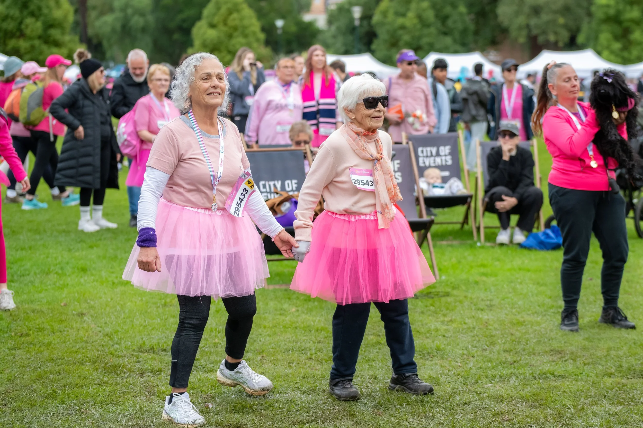 Two older racegoers wearing pink tutus hold hands and dance.