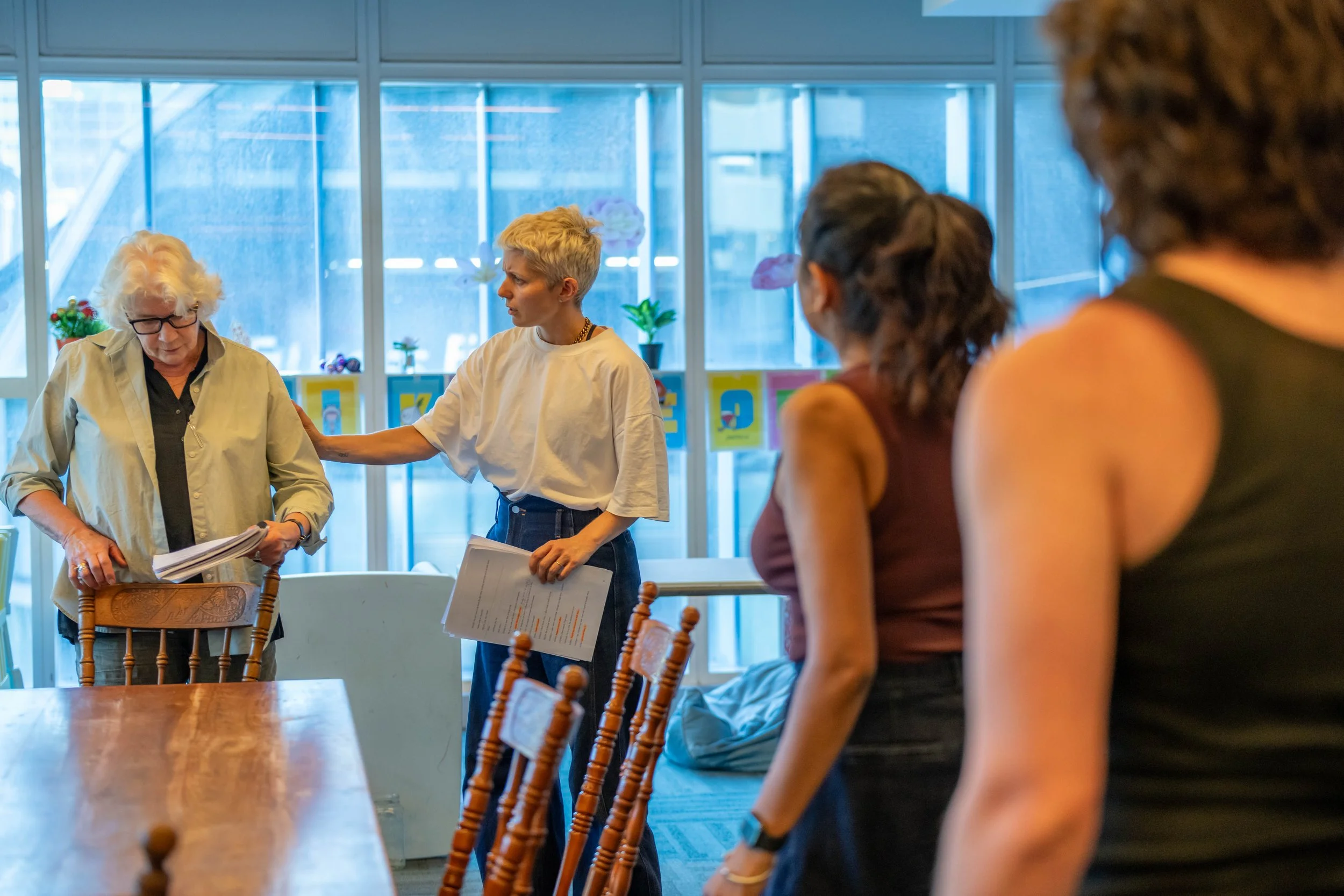 Four humans stand. One with short blonde hair places a hand tenderly on a human with white hair. The other two humans look on, their back is to the camera.