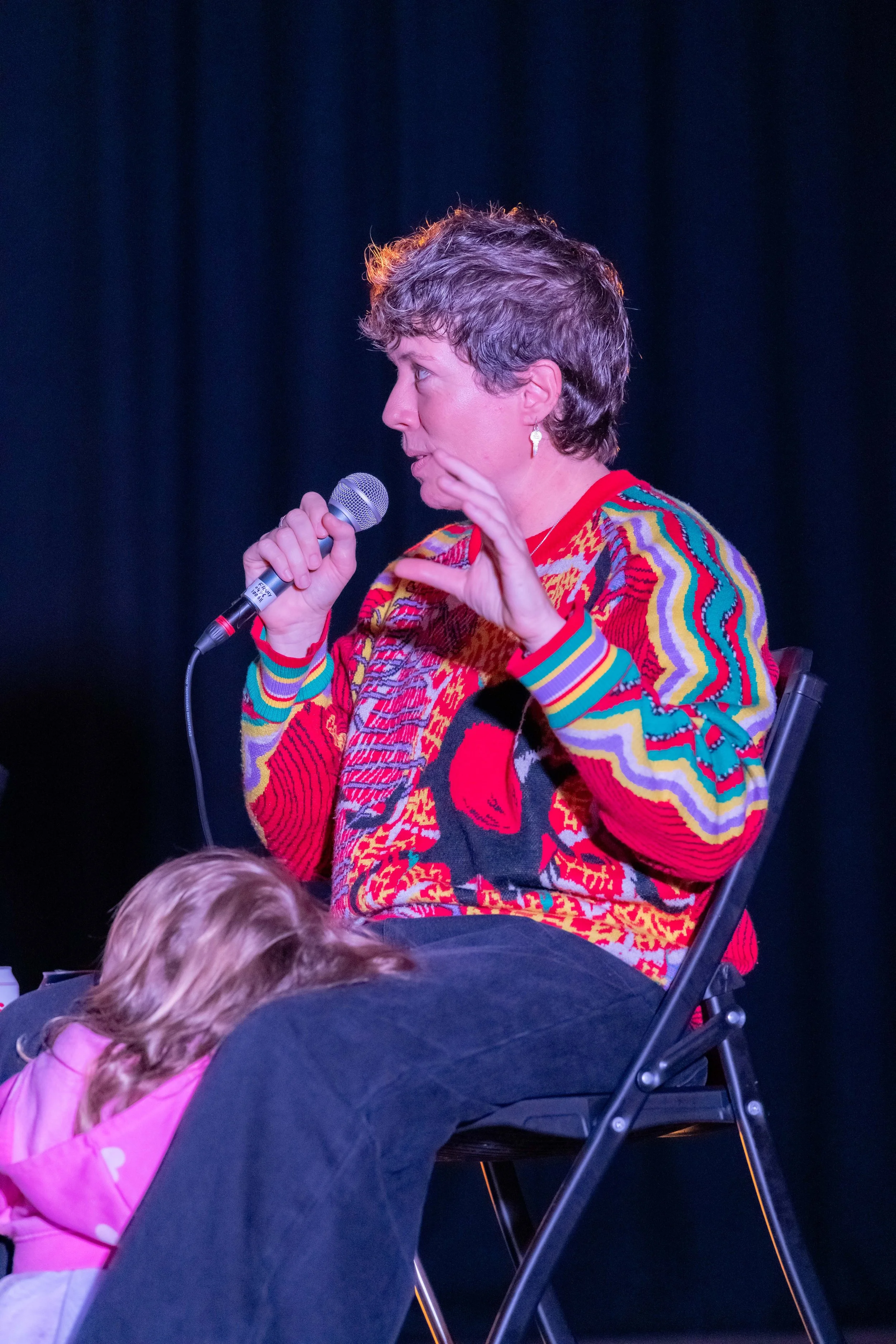 Mon Schafter speaks into a microphone whilst a small child rests between legs.