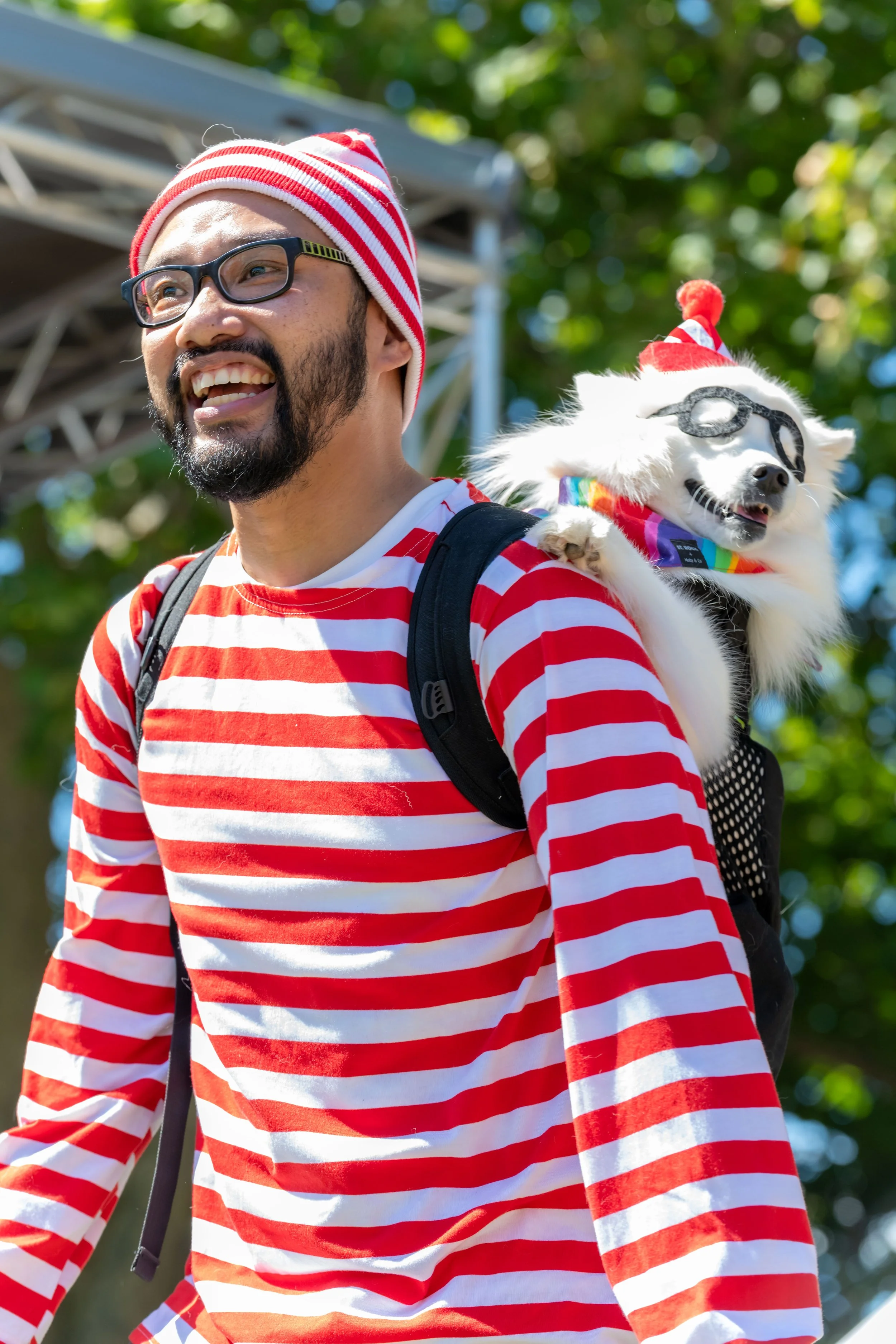 A human dressed as Waldo, from the Where's Wally books. In their backpack a white dog dressed as Woof sticks out.