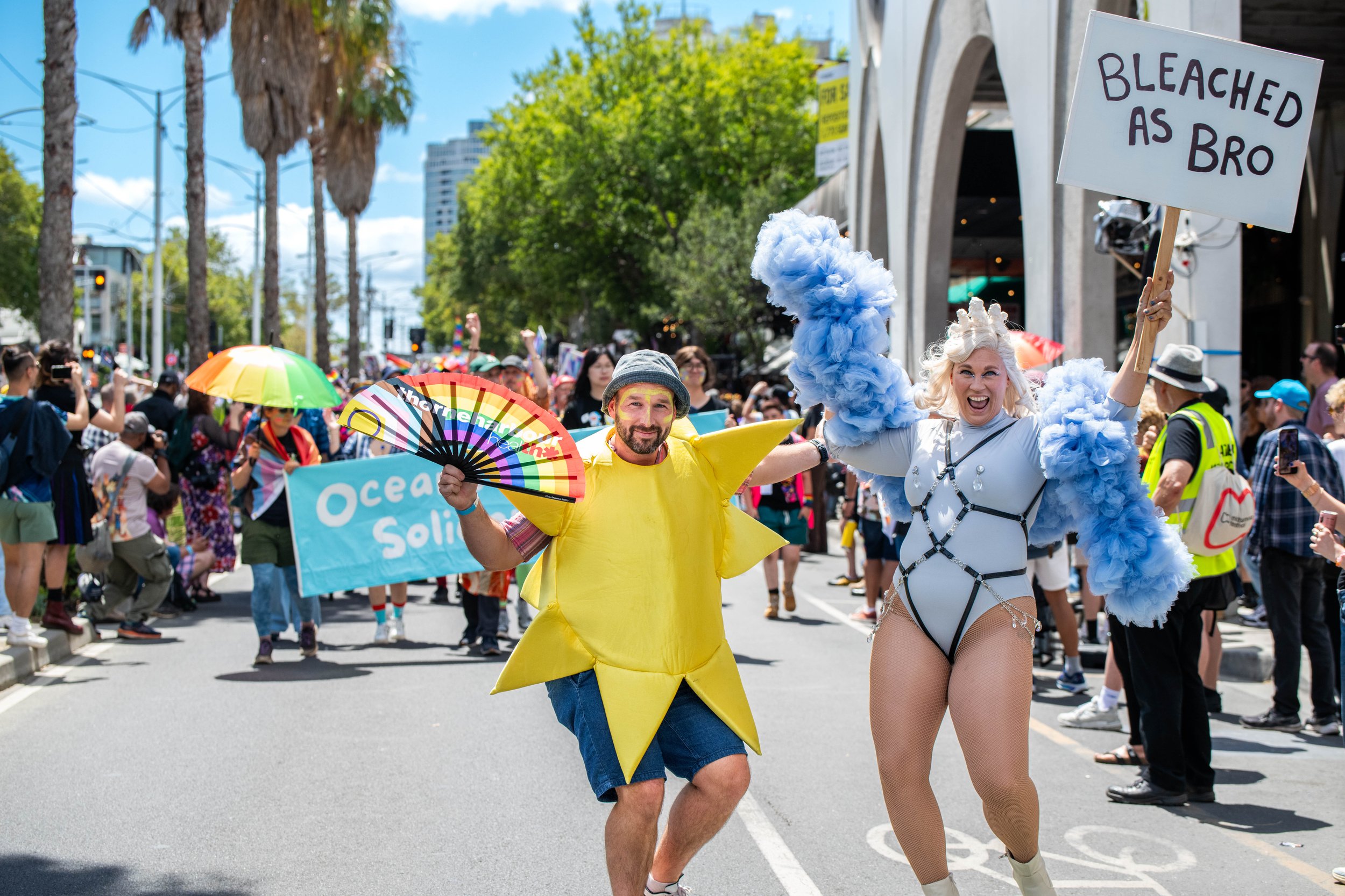 Two humans in costume pose for the camera. One human holds a placard up high "Bleached as bro".