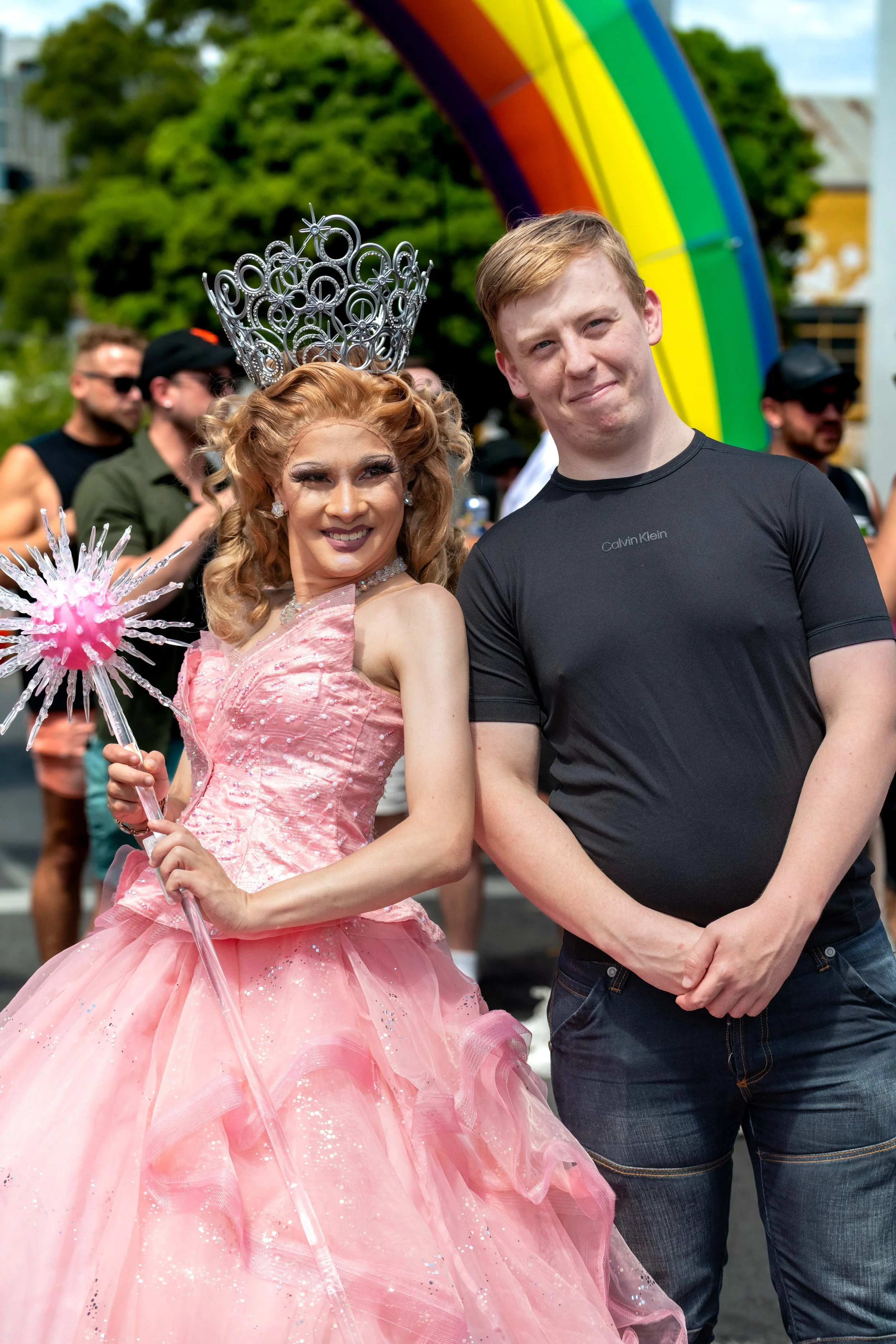 A drag queen princess poses with a human wearing black in front of a large inflattable rainbow.