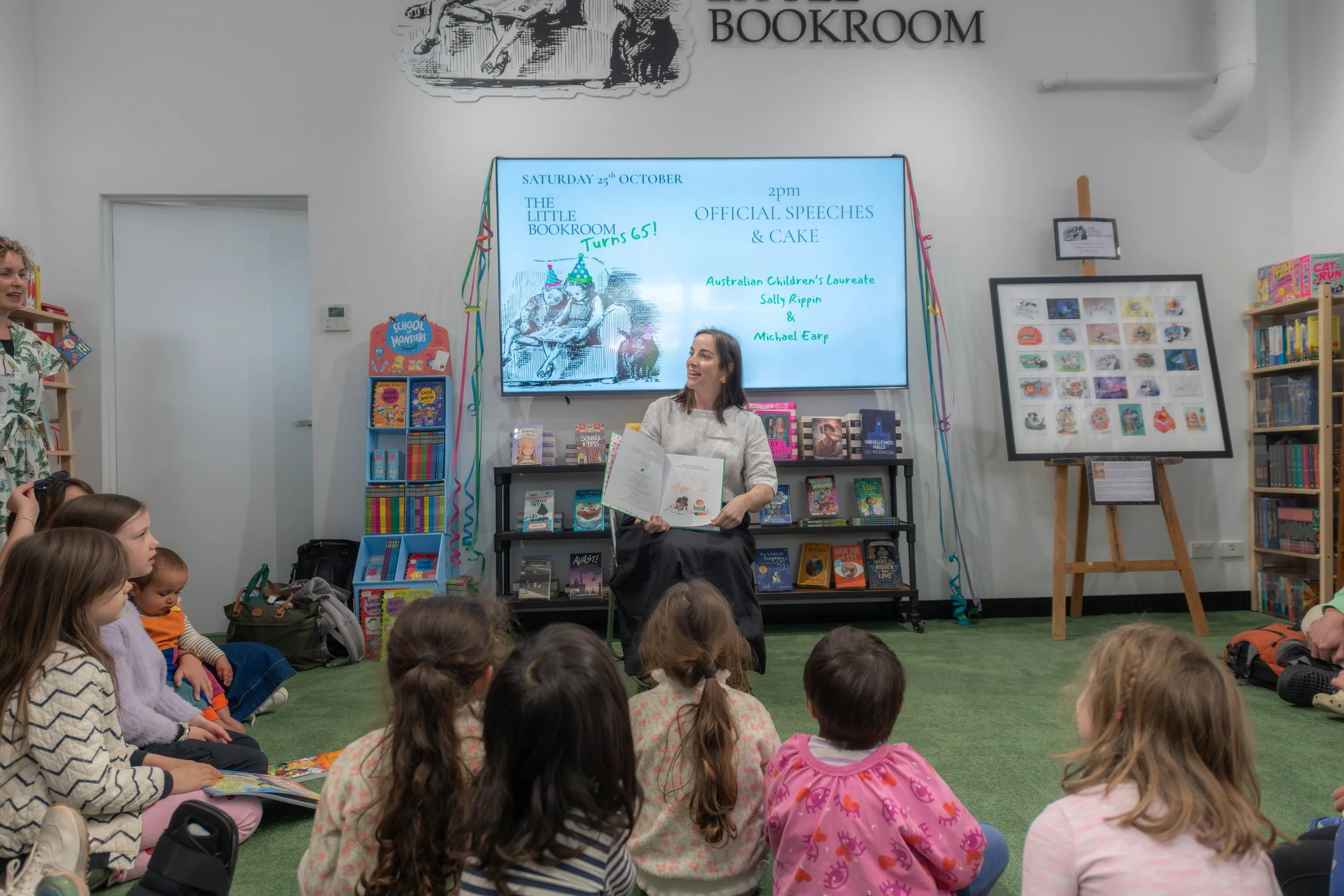 An author sits giving storytime to a group of children sitting on green carpet.