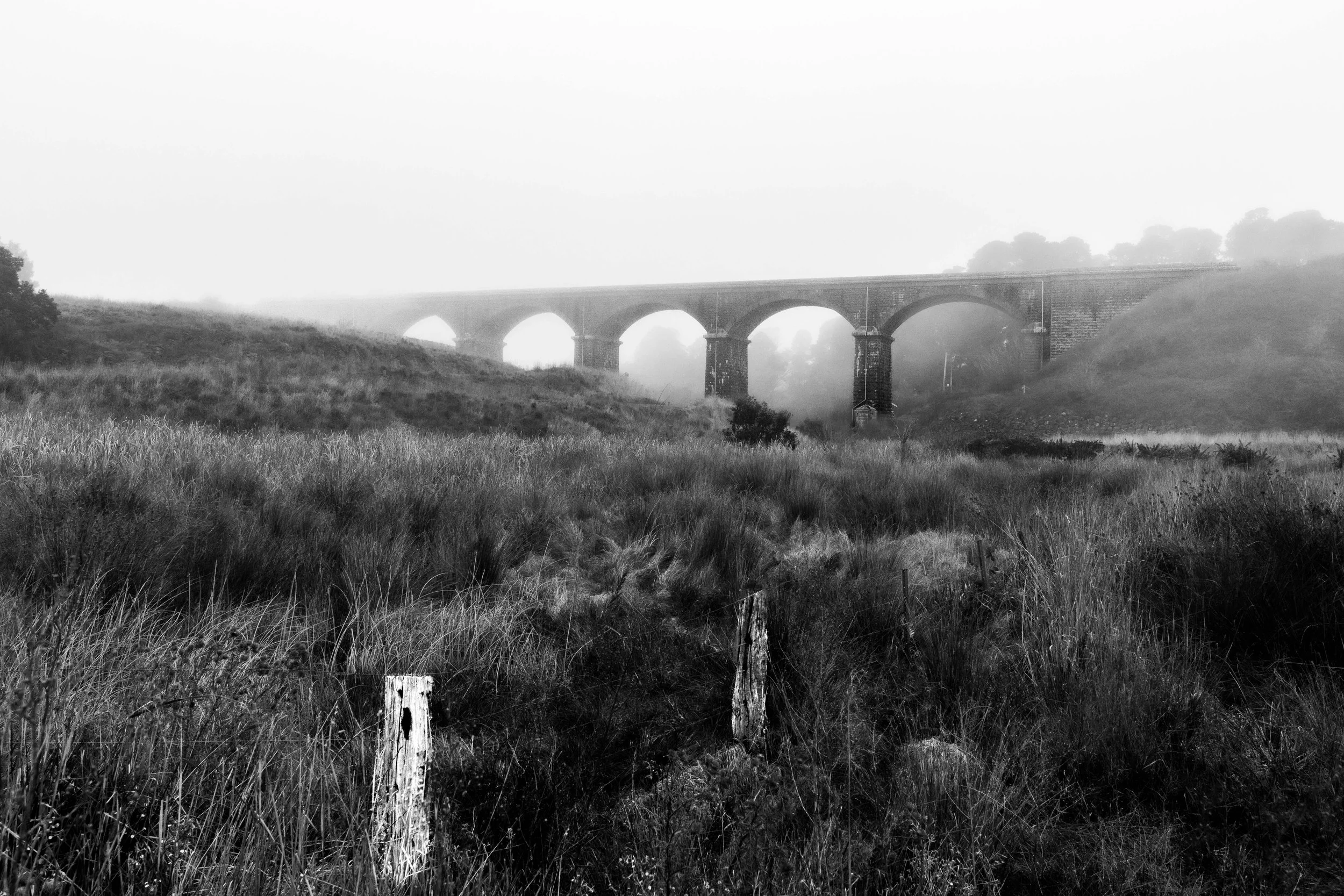 BNW landscape image of a brick viaduct through the thick fog. There is an old wooden and barbed wire fence in the foreground.