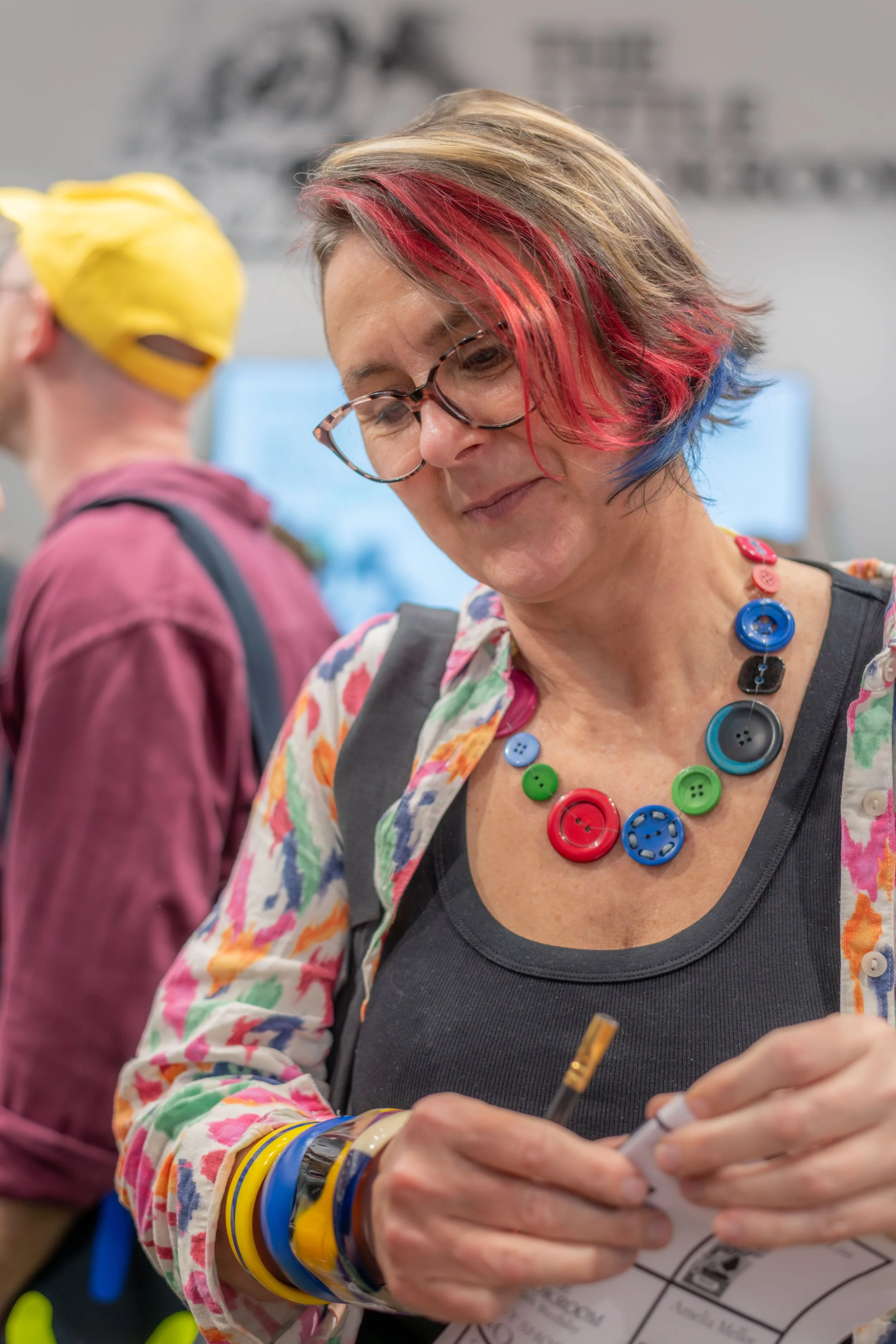 An author wearing a colourful necklace is folding paper and holding a pencil.