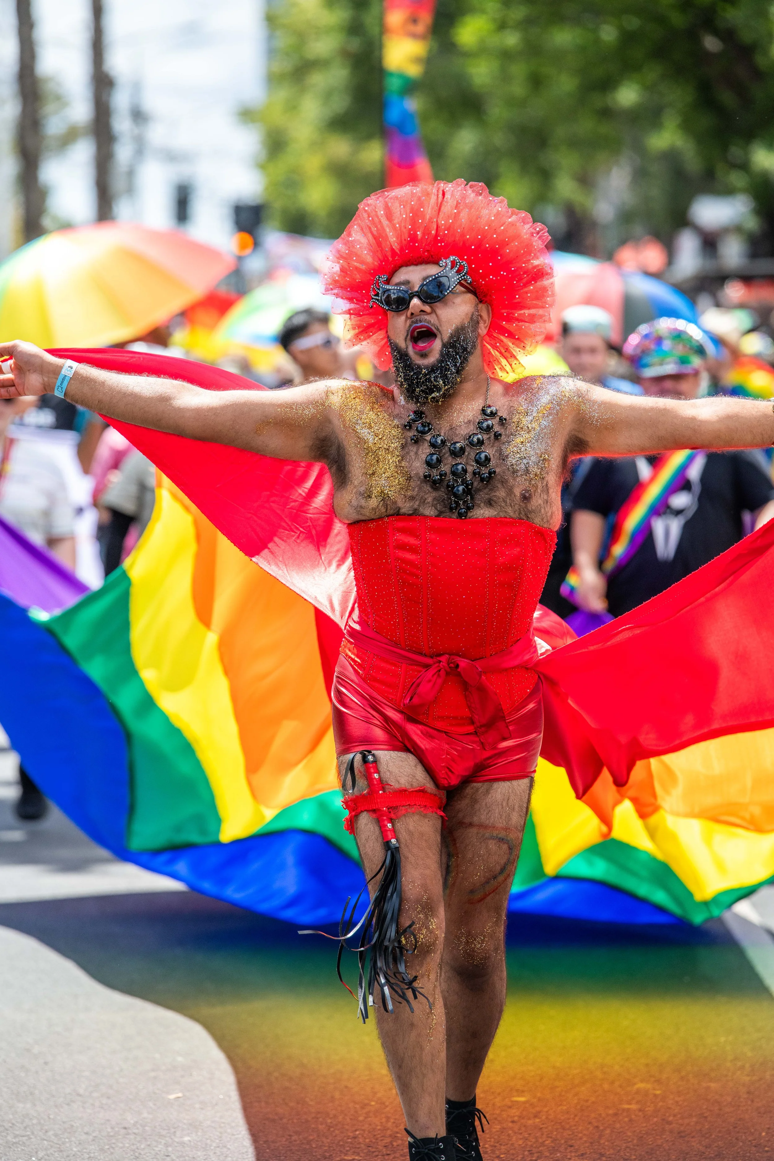 A human in red corset has a large pride flag trailling behind them.