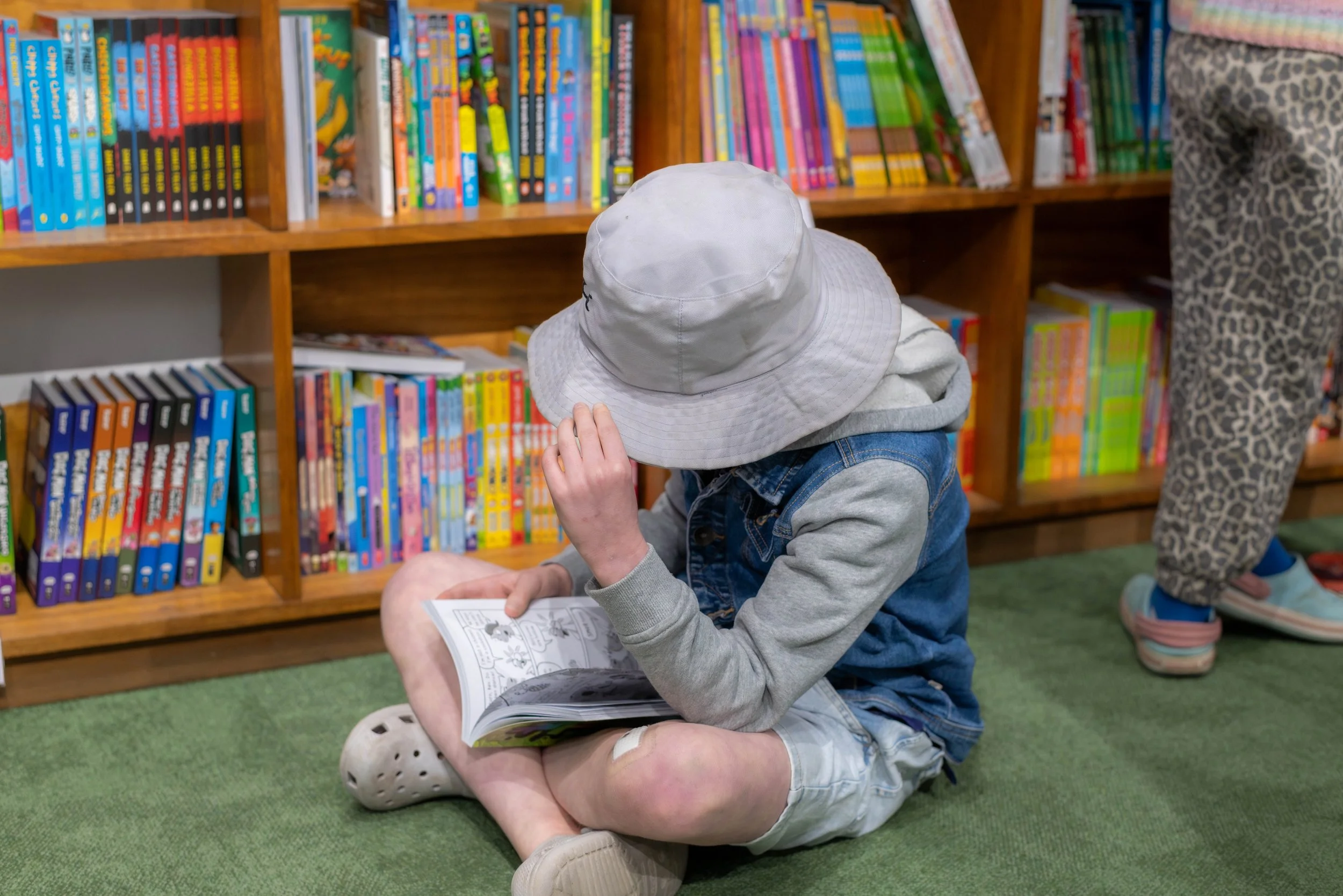A child in a grey bucket hat sits cross legged on the green carpet whilst reading a book.