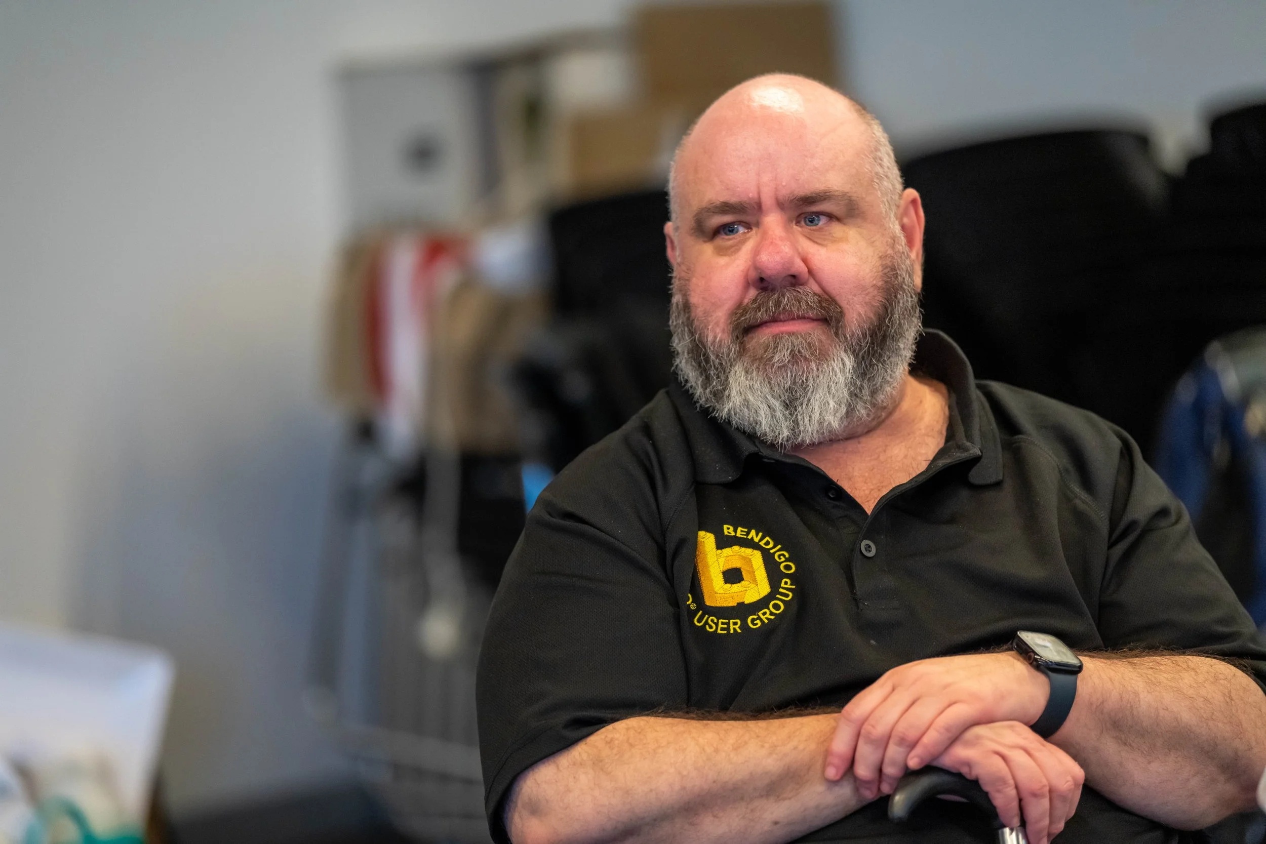 Ben MacEllen sits in a black polo holding onto a walking stick. He looks off to the side.