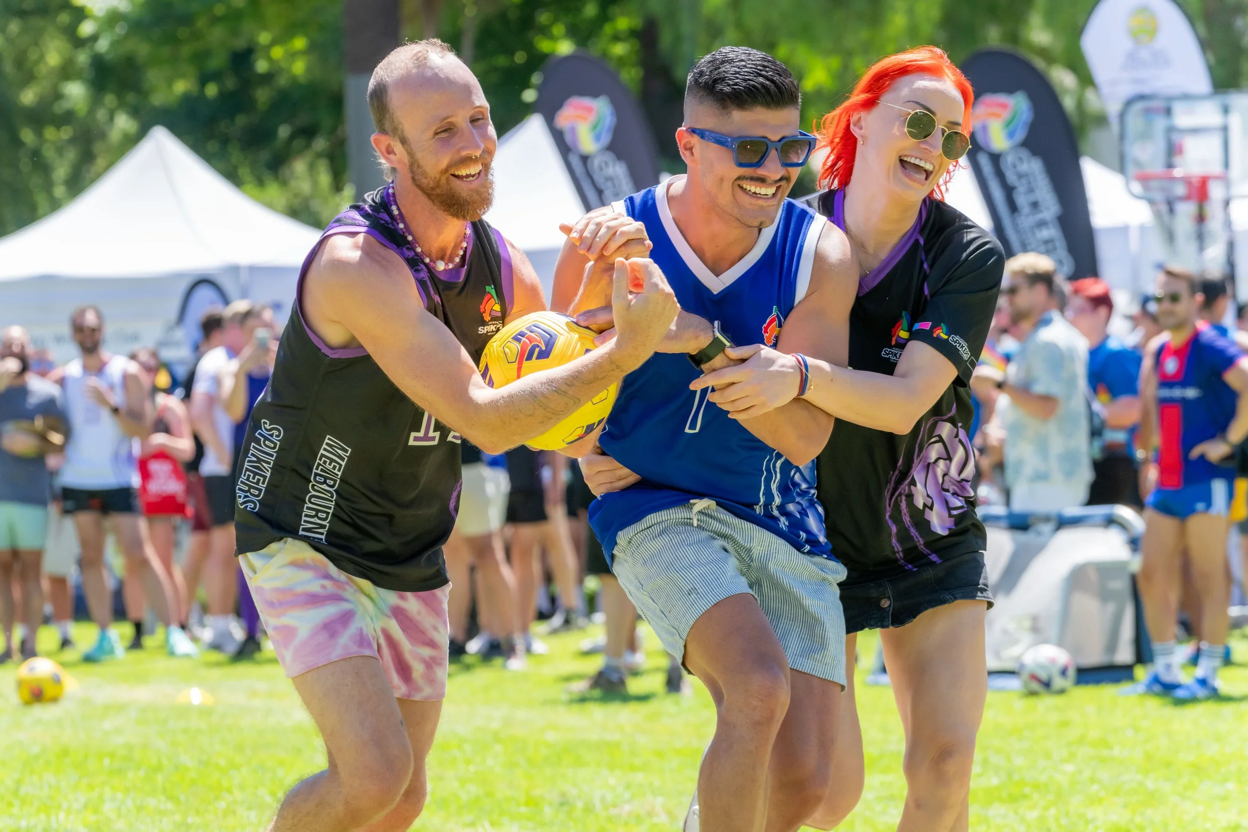 Three humans run along a field carrying a ball without using their hands. This is part of the Queerlympics.