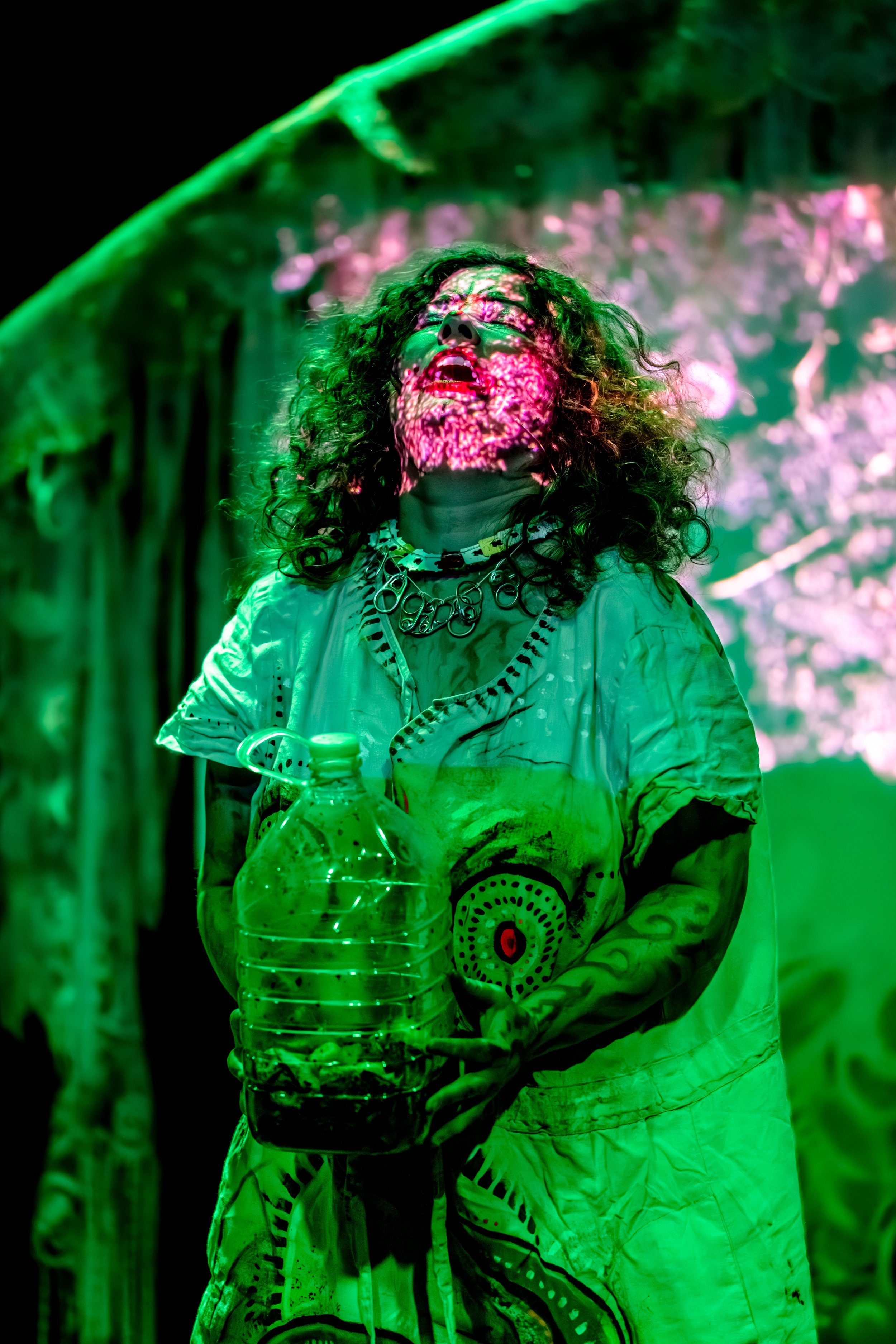 Jessie Ngaio holds a jug of worms. Jessie's face is lit pink as they stand in front of the screen. The rest of the staging is lit green.