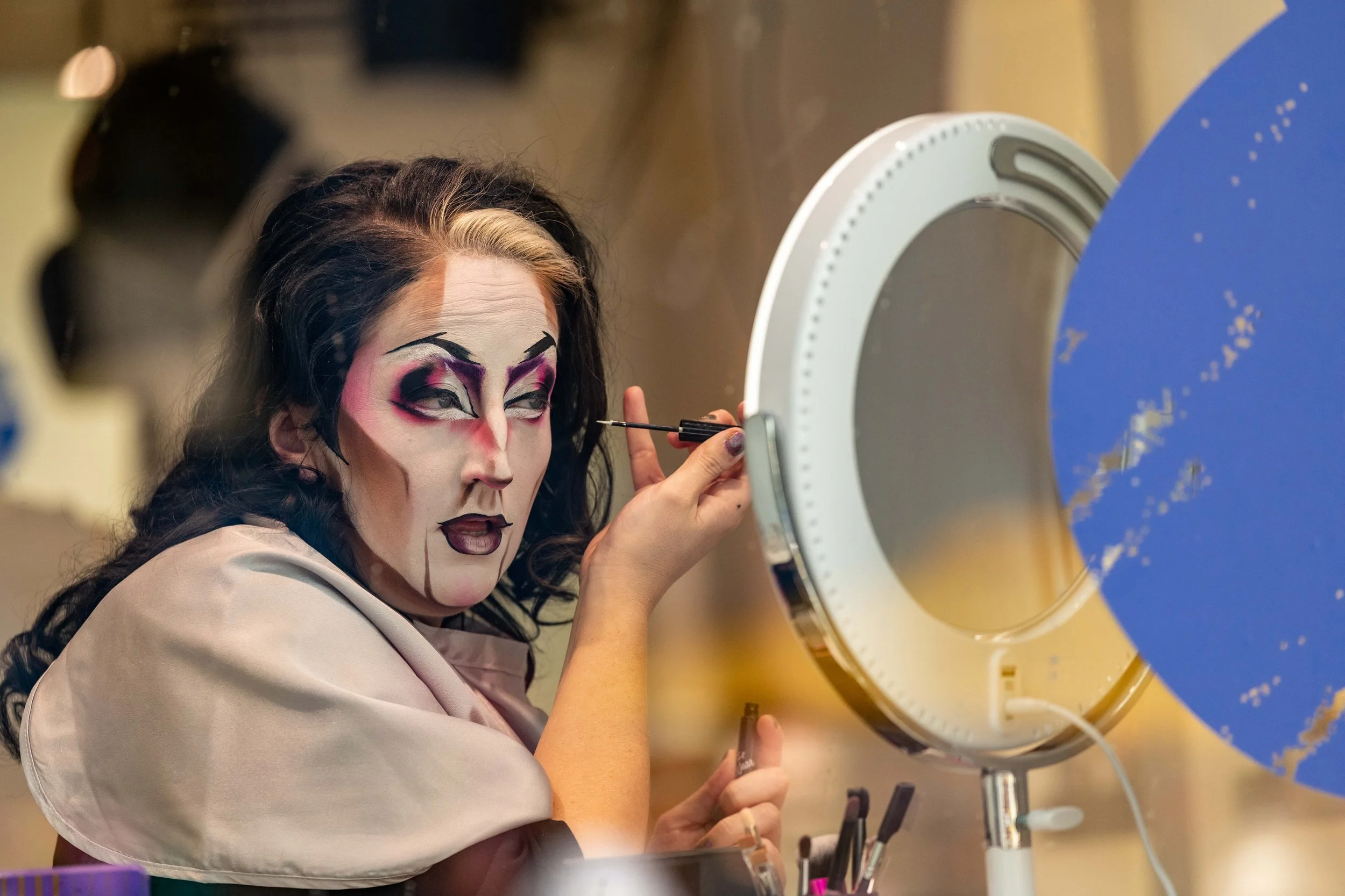 A drag king applies their face paint from behind a glass window.