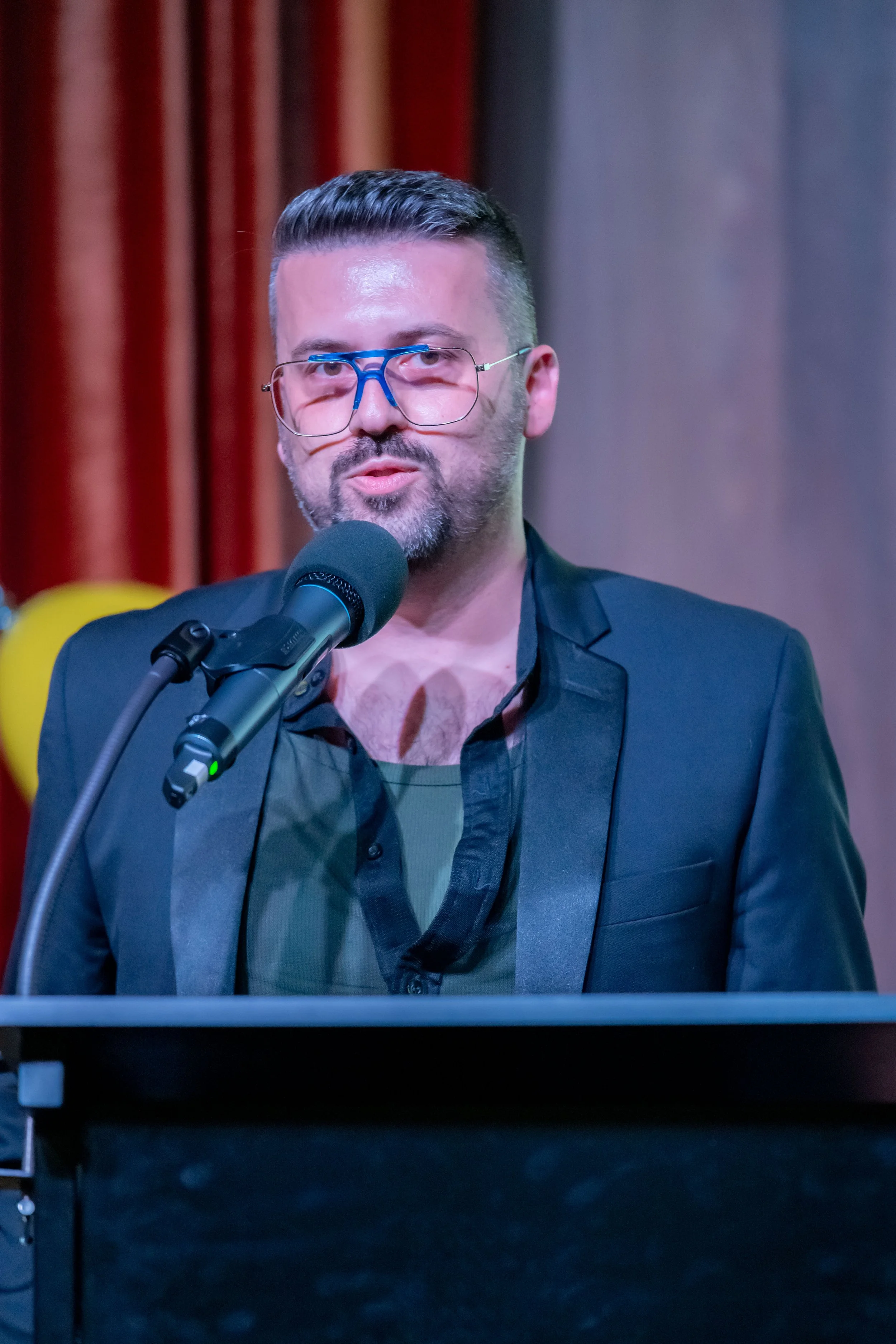 A human in a black dinner jacket stands at a lecturn and speaks into a microphone.