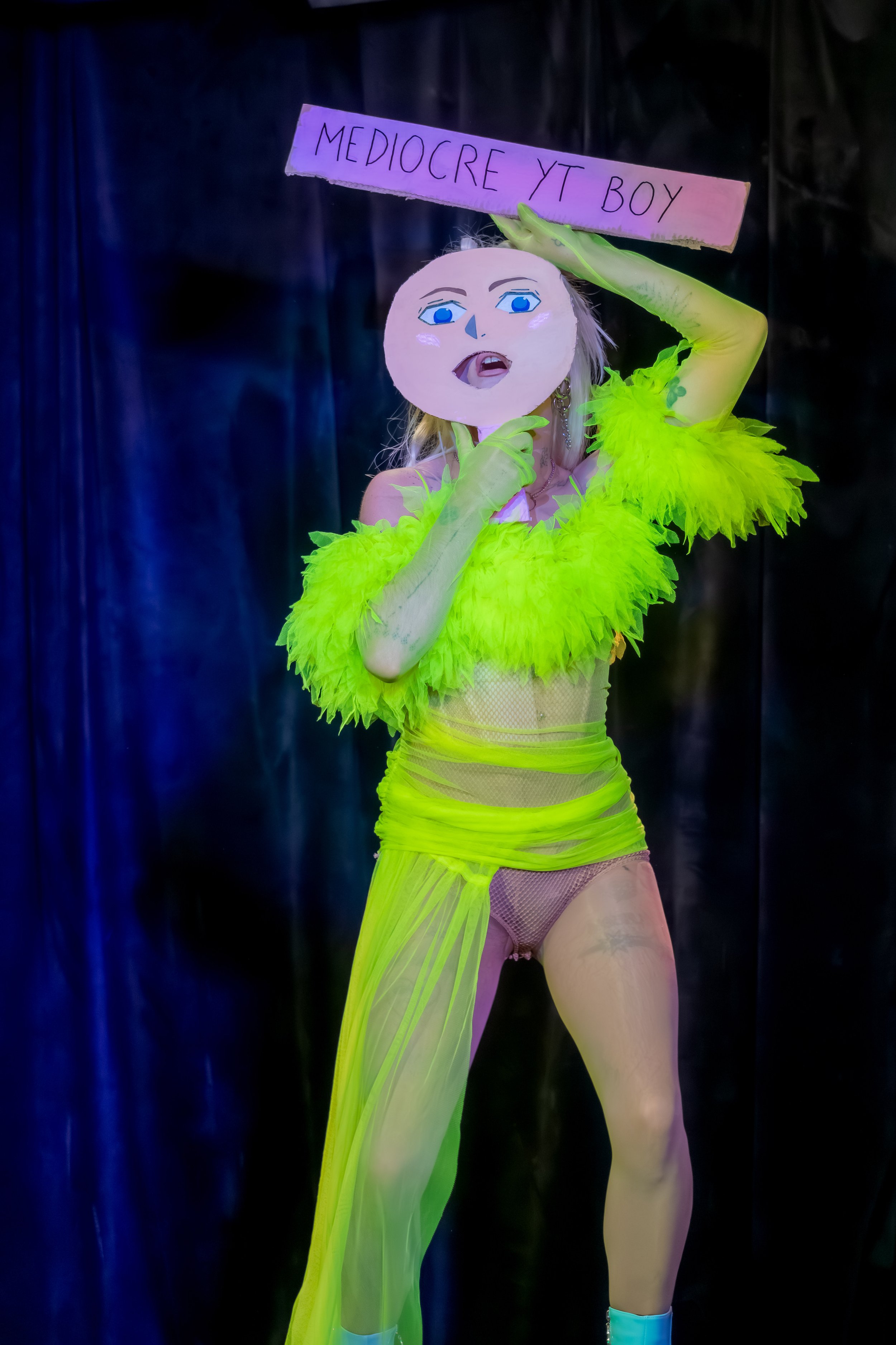 Mora Money, dressed in neon yellow, holds up a sign and a face mask. The sign says "Mediocre yt boy".