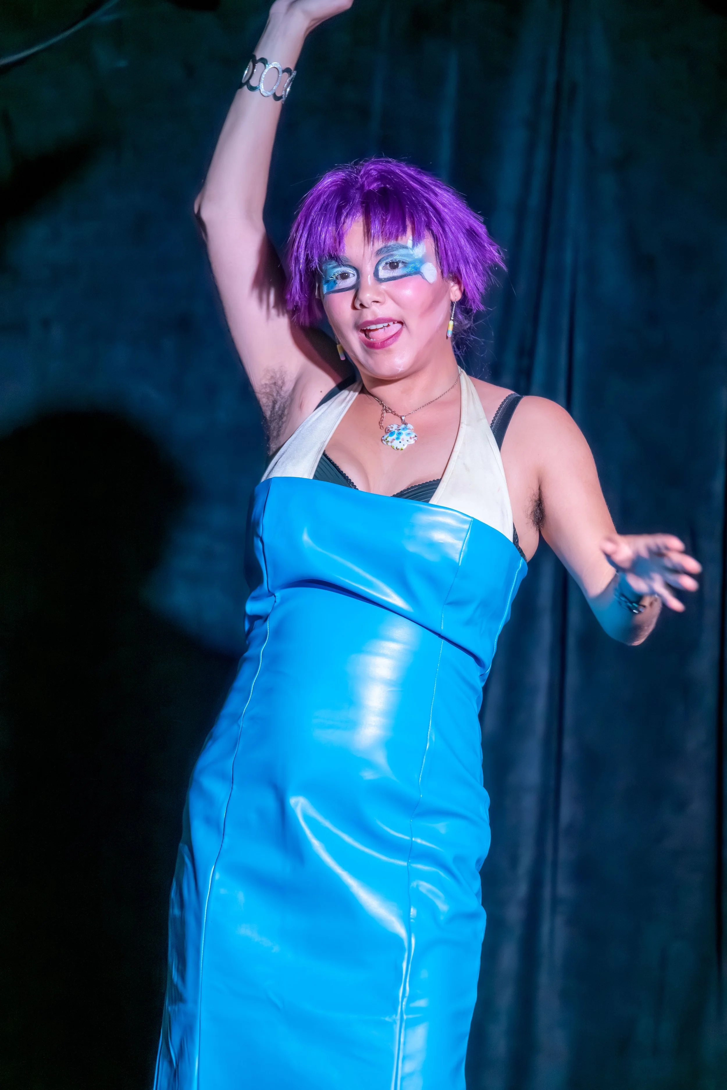 A drag queen in a purple wig and blue dress dances across the stage.