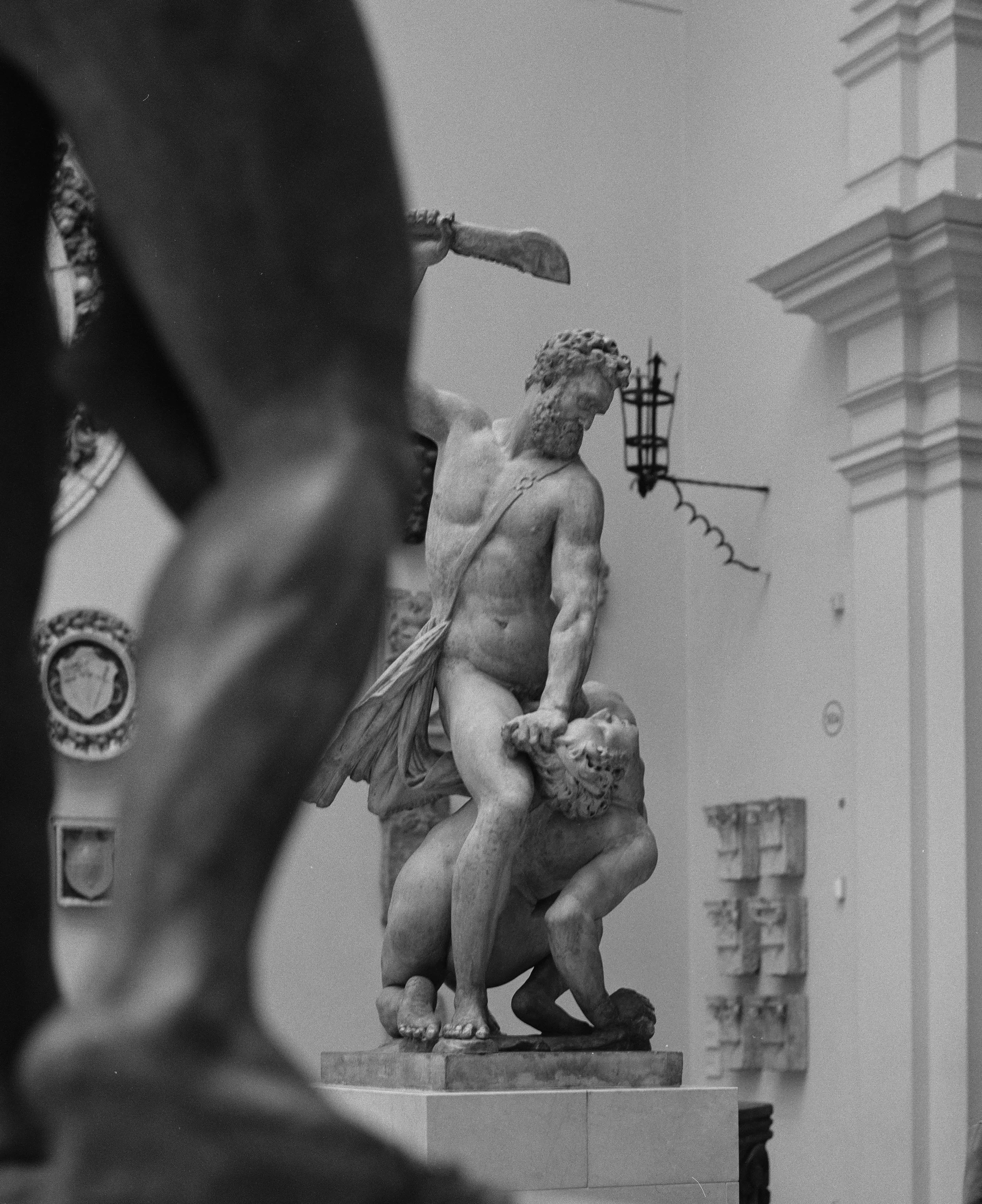 Black and white photo of an ancient marble sculpture depicting a mythological scene, with a muscular male figure engaging another figure, displayed indoors with architectural elements and decorative wall pieces.