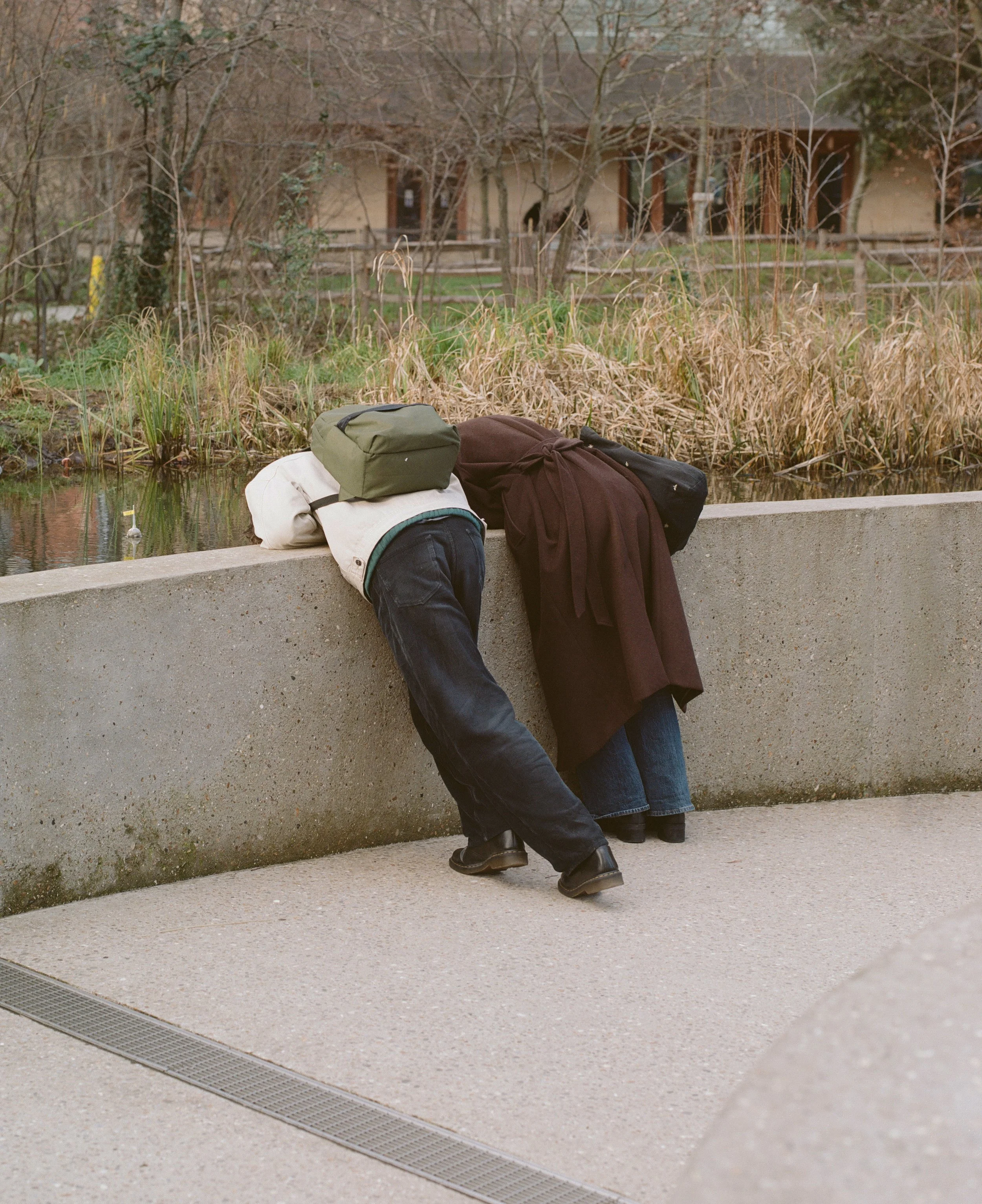Two people with backpacks leaning over a concrete ledge by a pond, with trees and a building in the background.