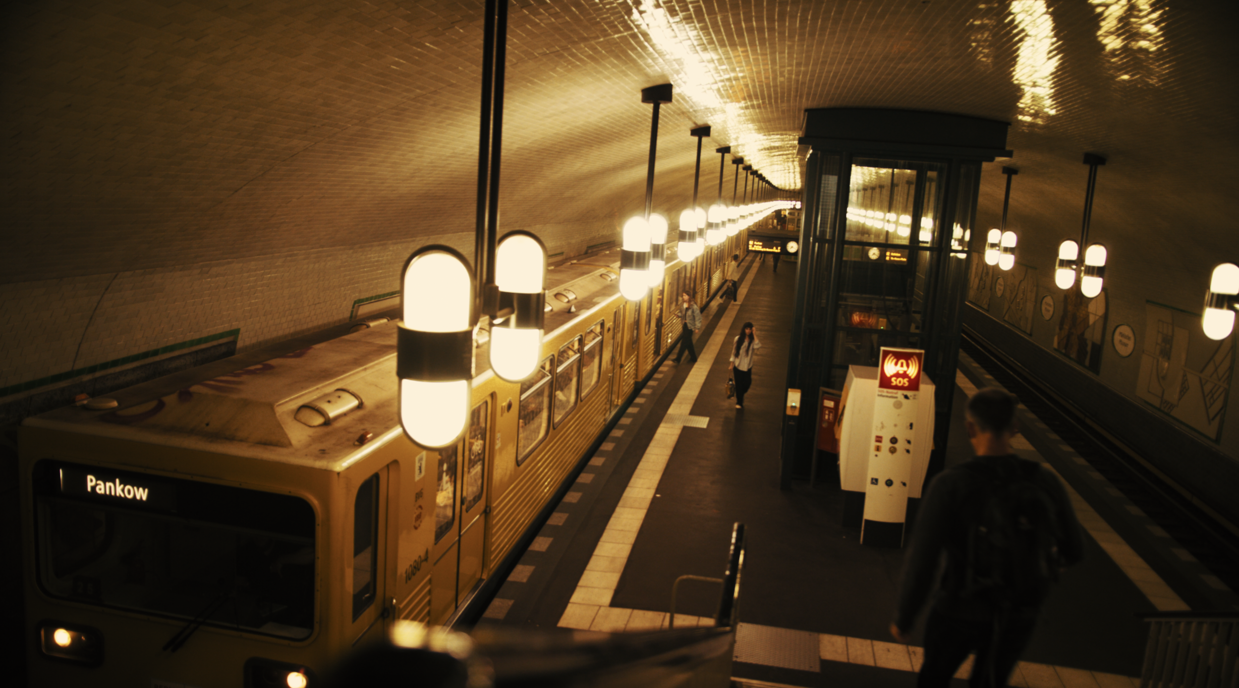 Underground train station with yellow train on tracks, people walking on platform, illuminated hanging lamps, and an elevator shaft. Sign indicates the destination "Pankow."