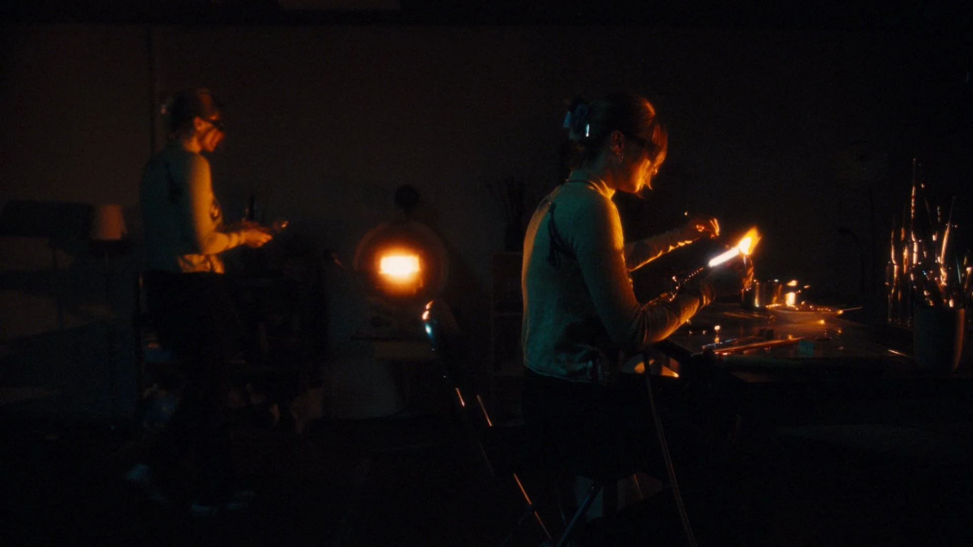 Two women working at a jewelry or craft station in a dark room illuminated by fire or candle light, with one woman focused on her task and the other standing nearby.