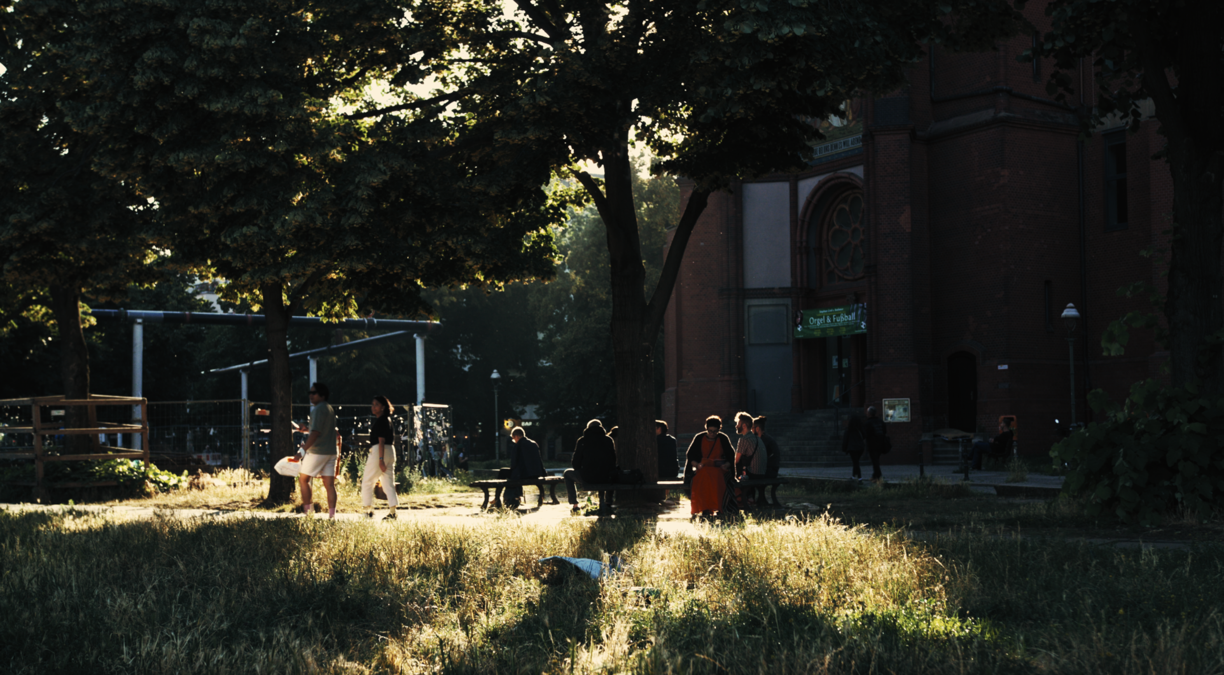 People walking and sitting in a park during late afternoon or early evening, with large trees and a brick building in the background.
