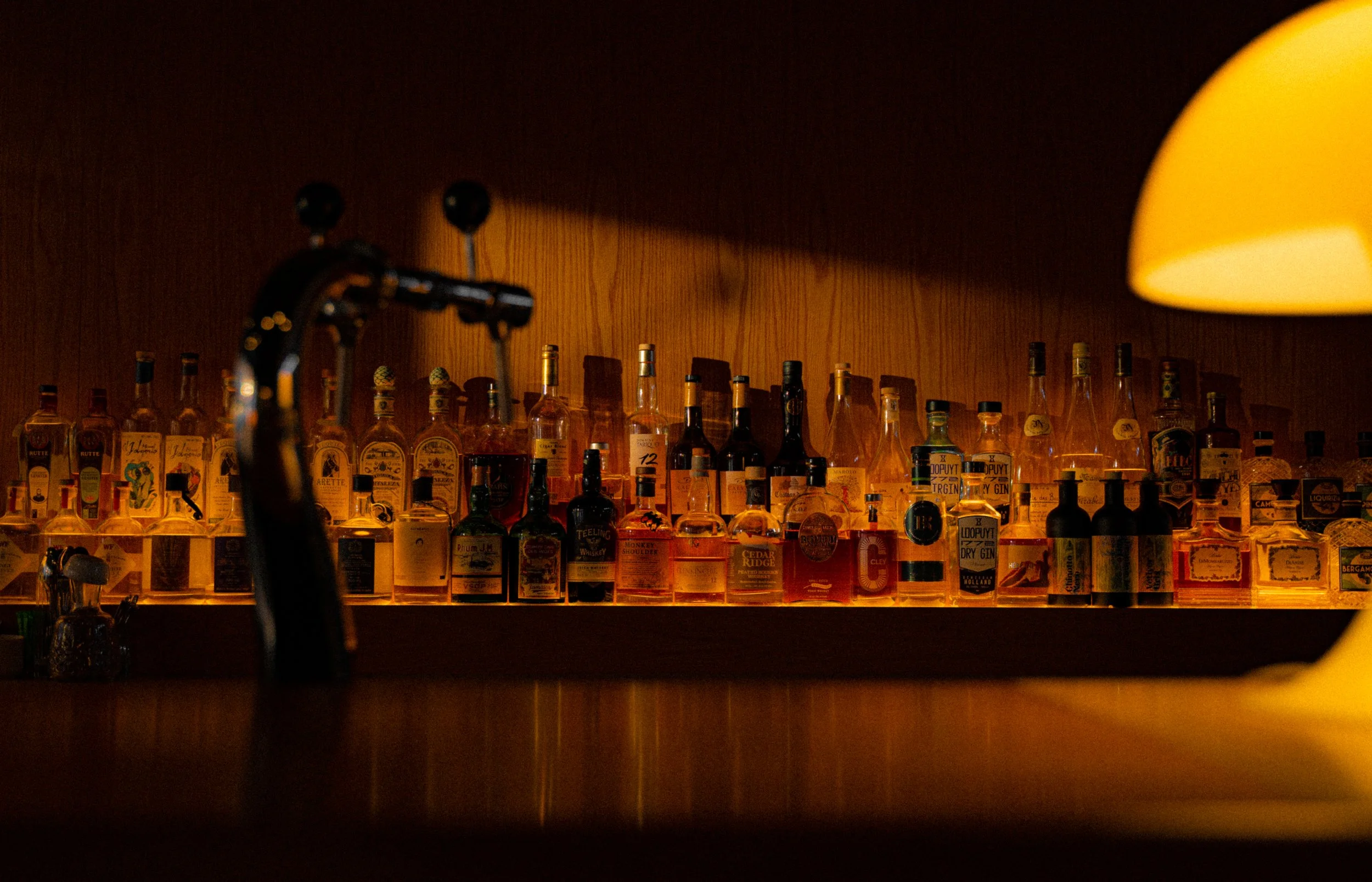 A bar shelf with a variety of liquor bottles and a yellow lamp on the right side.