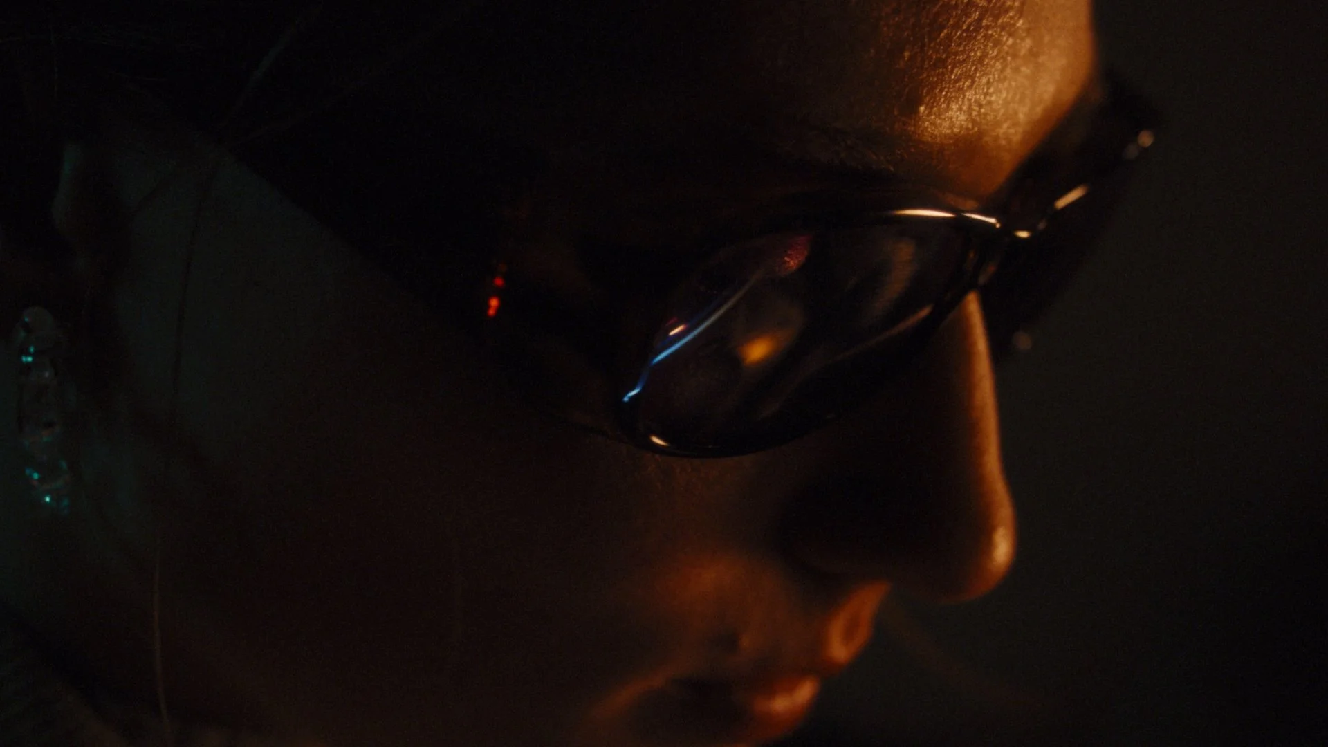 Close-up of a person's face in darkness with glasses reflecting faint light, showing a nose and lips.