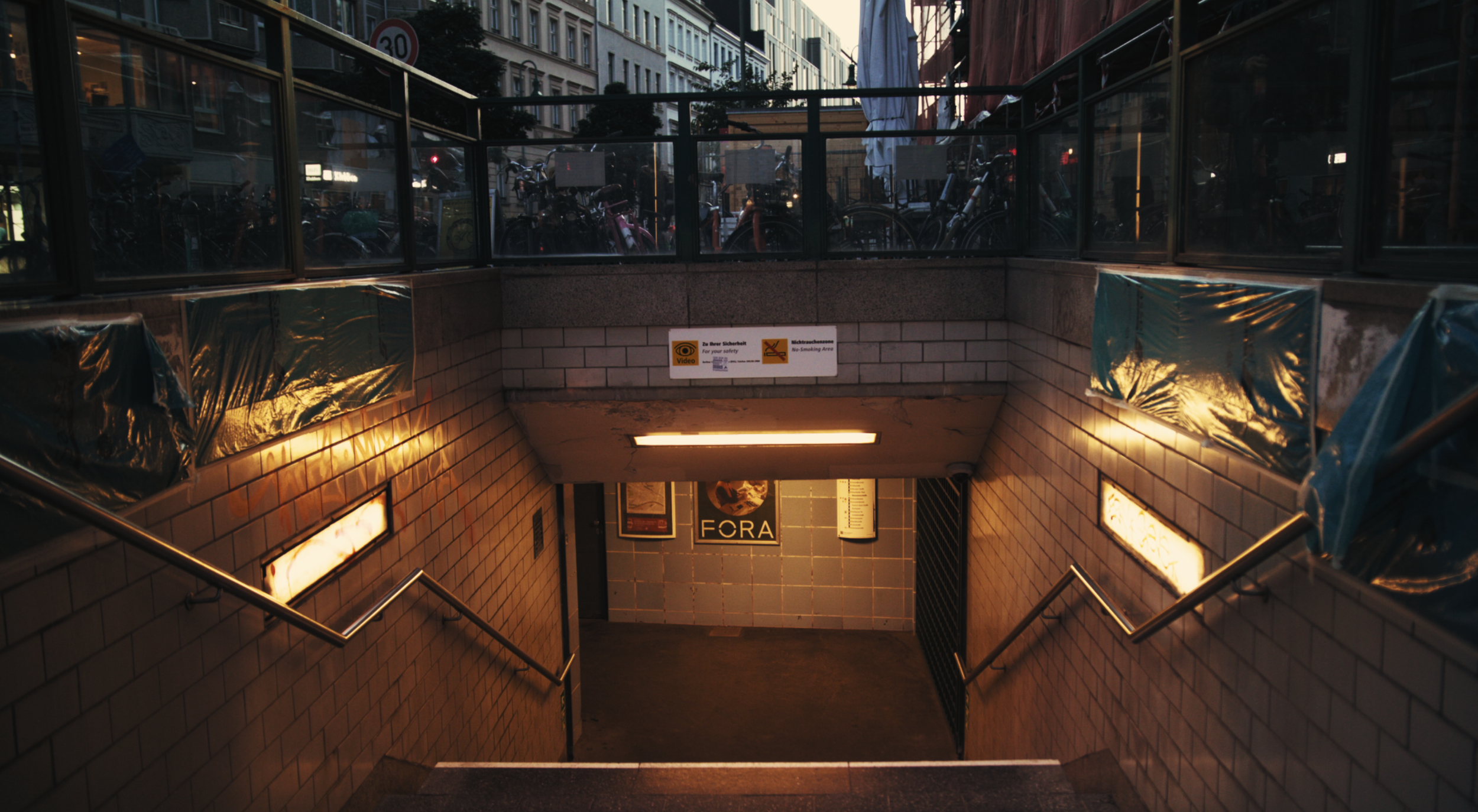 Staircase leading underground with tiled walls, metal handrails, and signage, in an urban setting.
