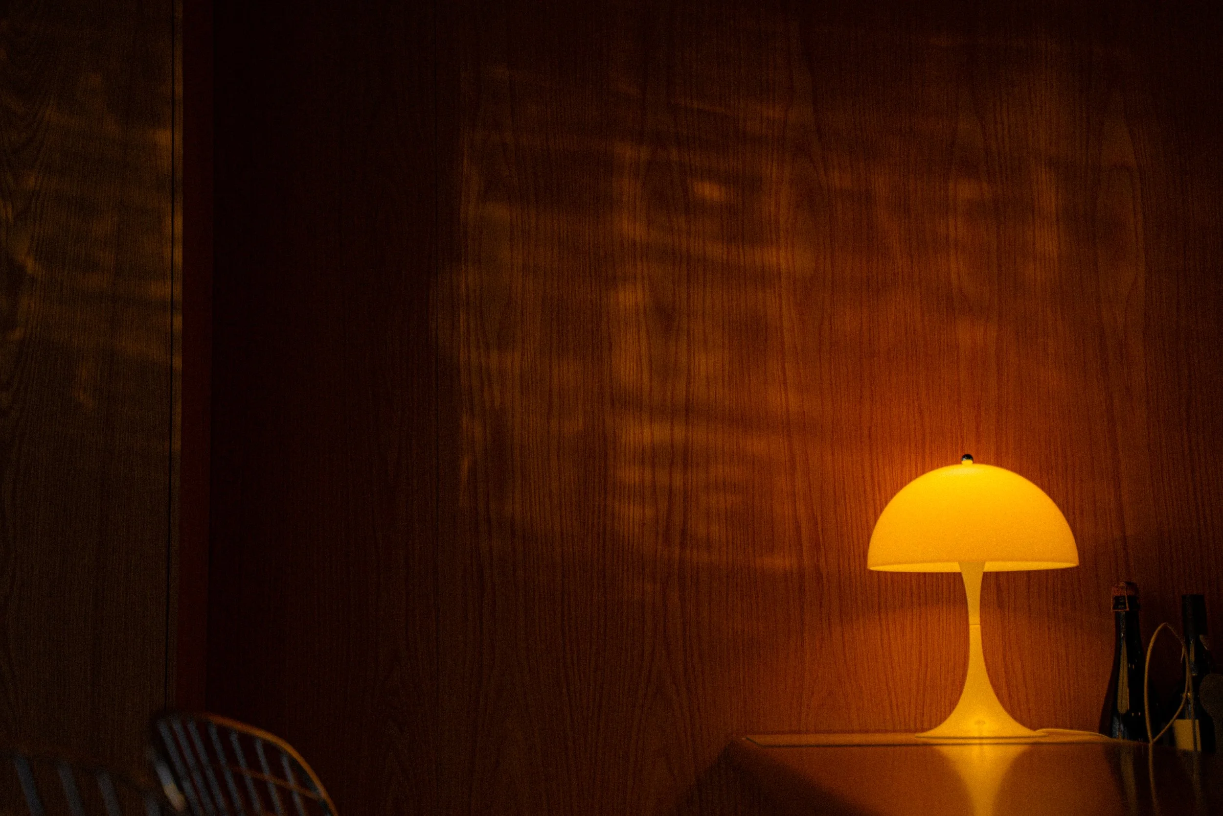 A yellow table lamp with a rounded shade on a wooden surface, next to an electrical outlet, with a dark wooden wall in the background.