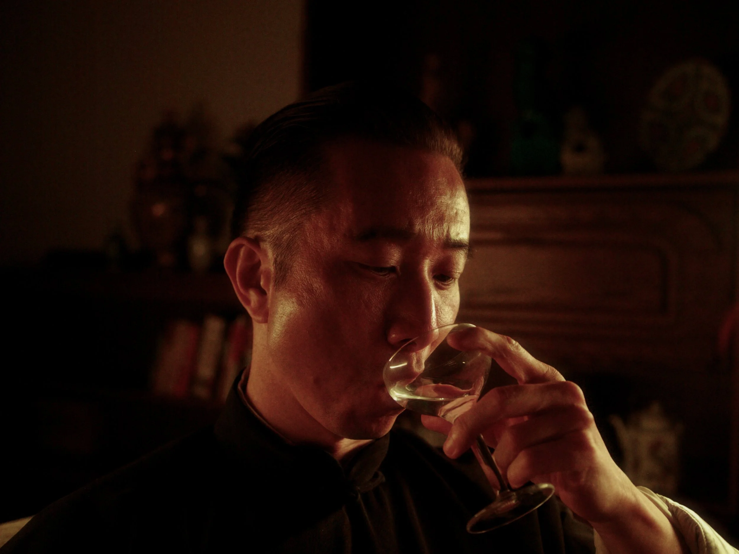 A person with short dark hair and a side part is drinking from a wine glass in a dimly lit room.