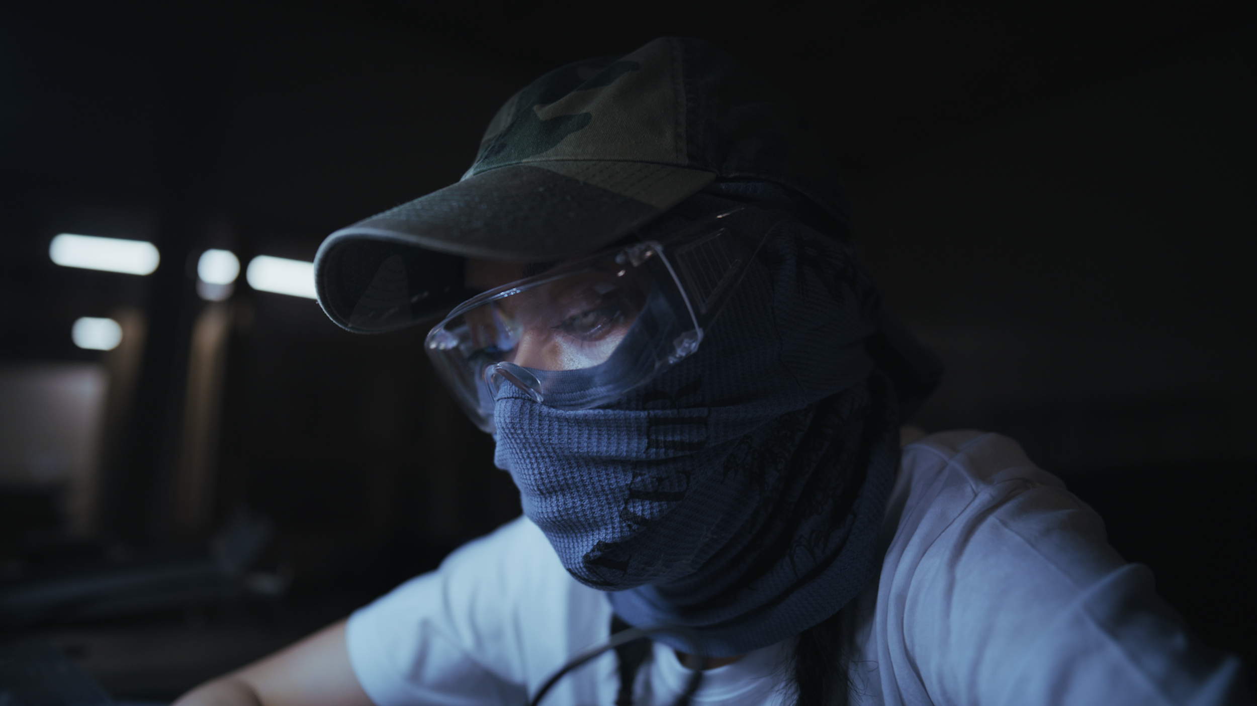 Person wearing camouflage cap, protective glasses, and a face covering, working in a dimly lit environment.