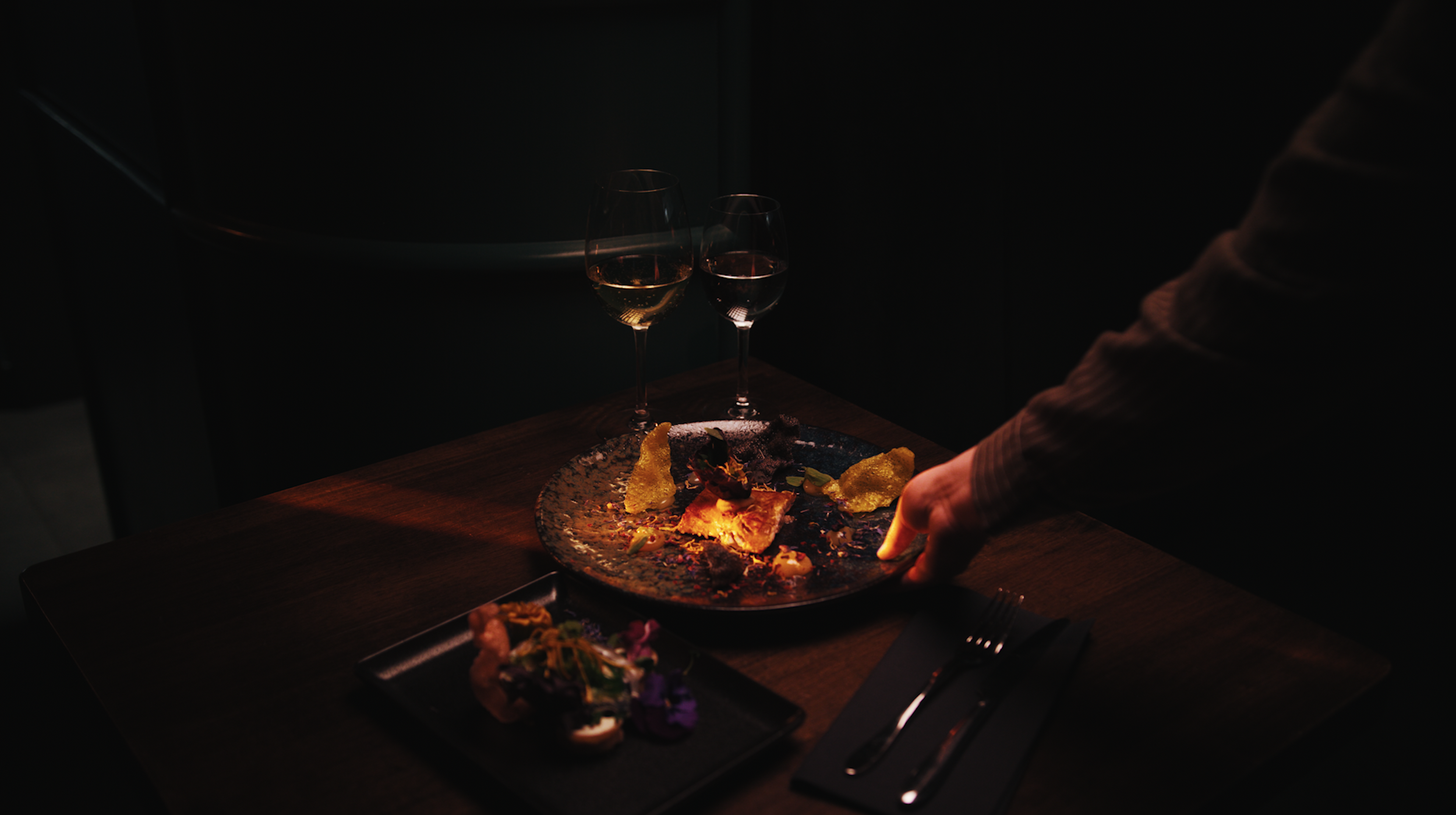 A dimly lit restaurant table with two glasses of white wine and a plated dessert with decorative garnishes.