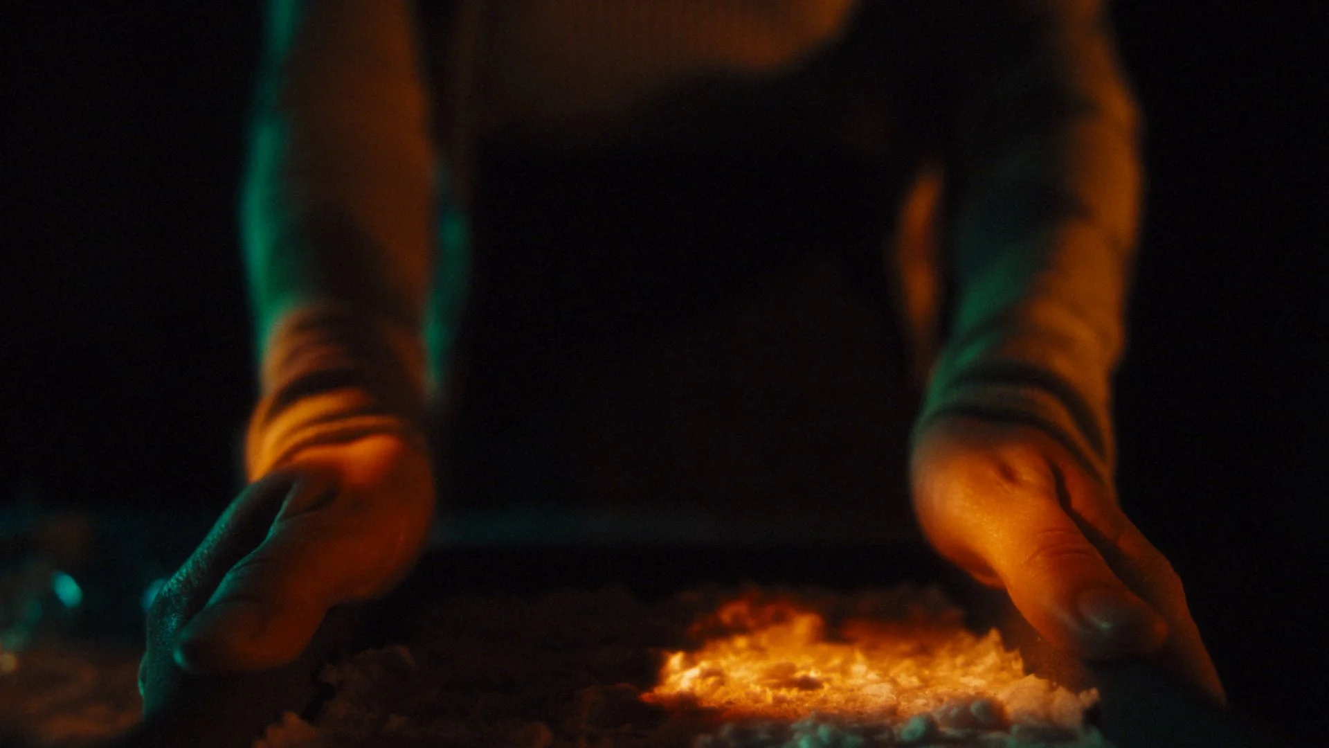 Person with glowing hands crouching over a fire in darkness, illuminated by orange flame and subtle teal lighting.