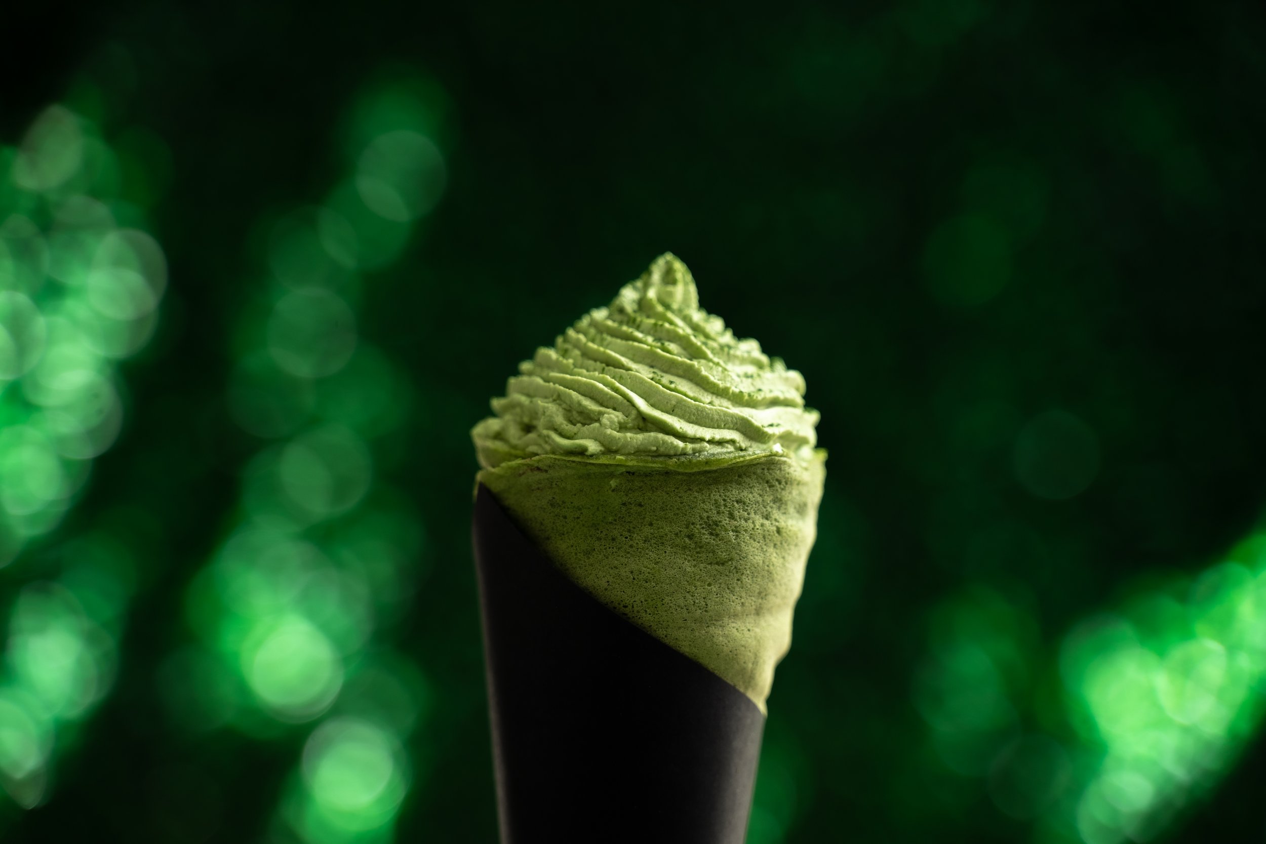 Close-up of a vanilla ice cream cone topped with green-colored whipped cream or frosting, against a dark background with circular green bokeh lights.
