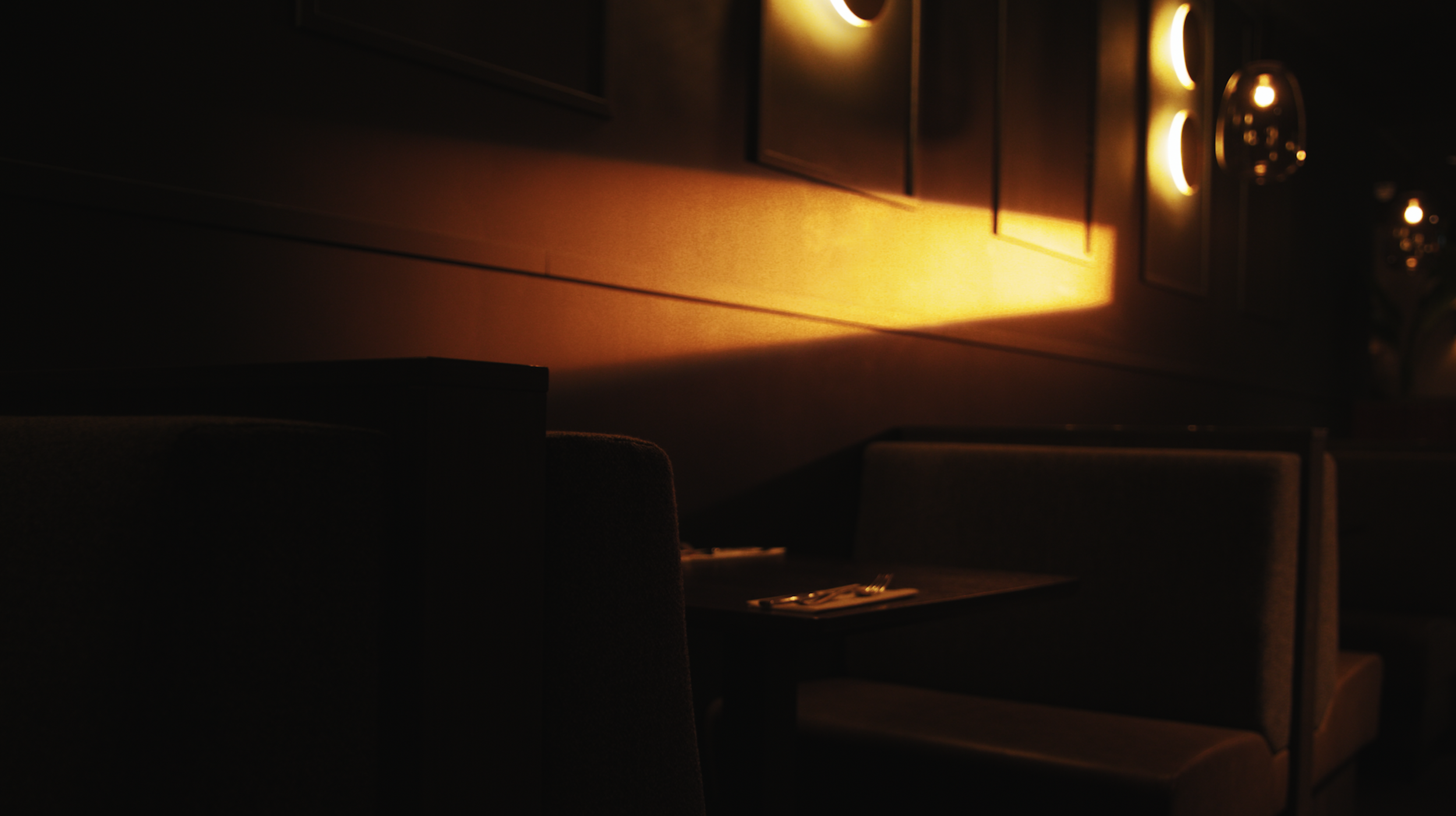 Dimly lit dining room with warm yellow lighting, a table set with utensils, and a dark wall with modern light fixtures