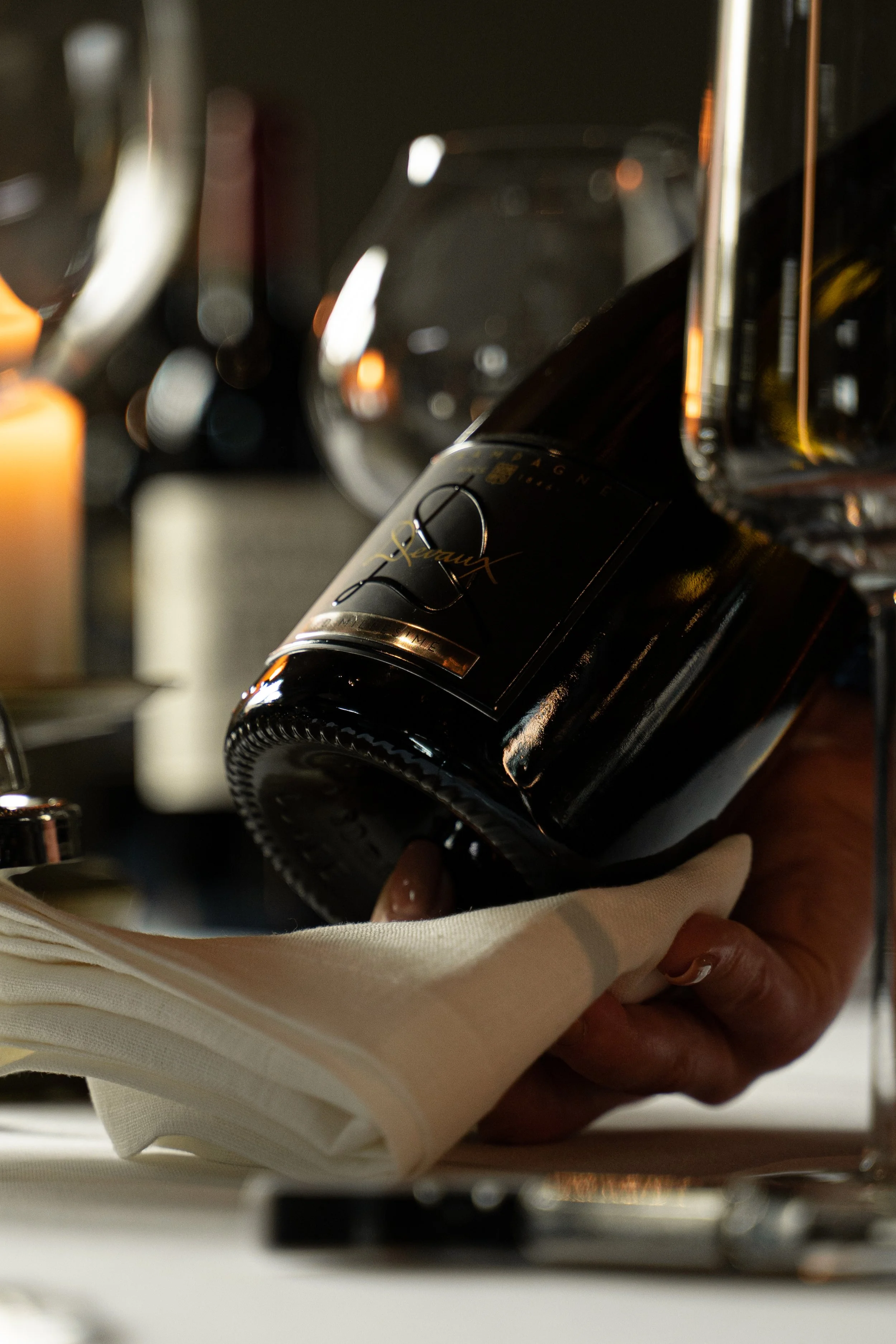 A hand holding a black bottle with gold accents, labeled 'Xawes' in gold, with a silver cap, against a blurred background of hanging objects.