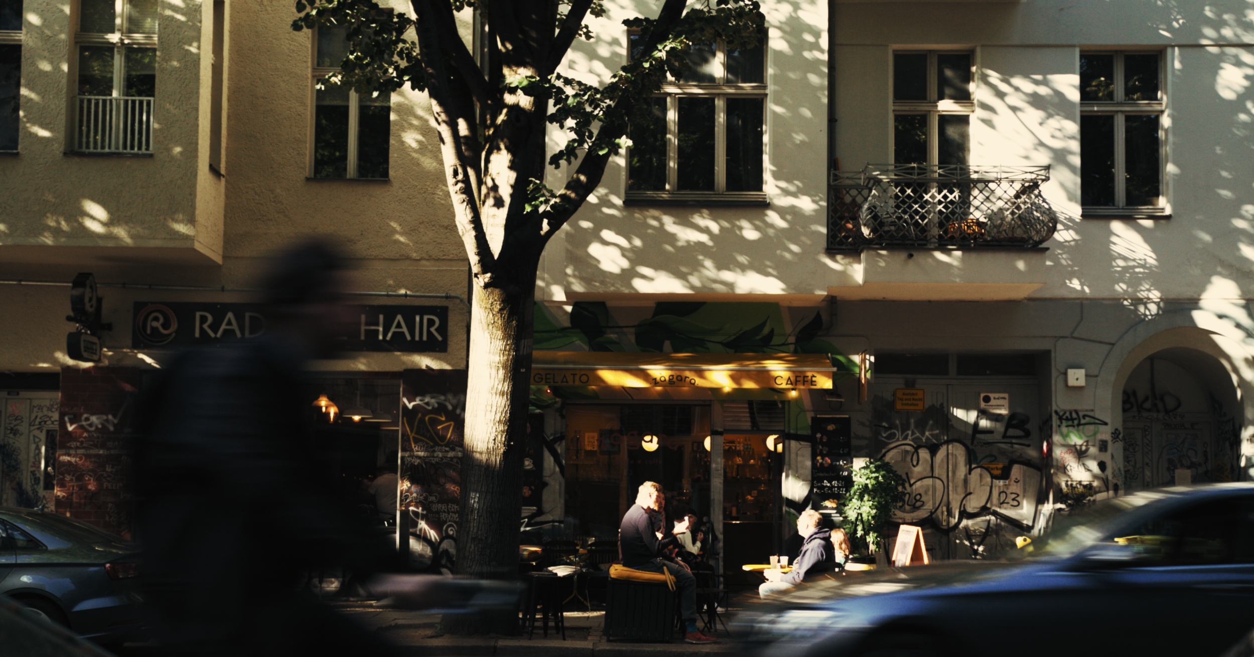 A city street scene with a building featuring a cafe on the ground level and residential windows above. The cafe has outdoor seating with people sitting and talking. A tree in front of the building casts shadows on the facade, and there is graffiti o
