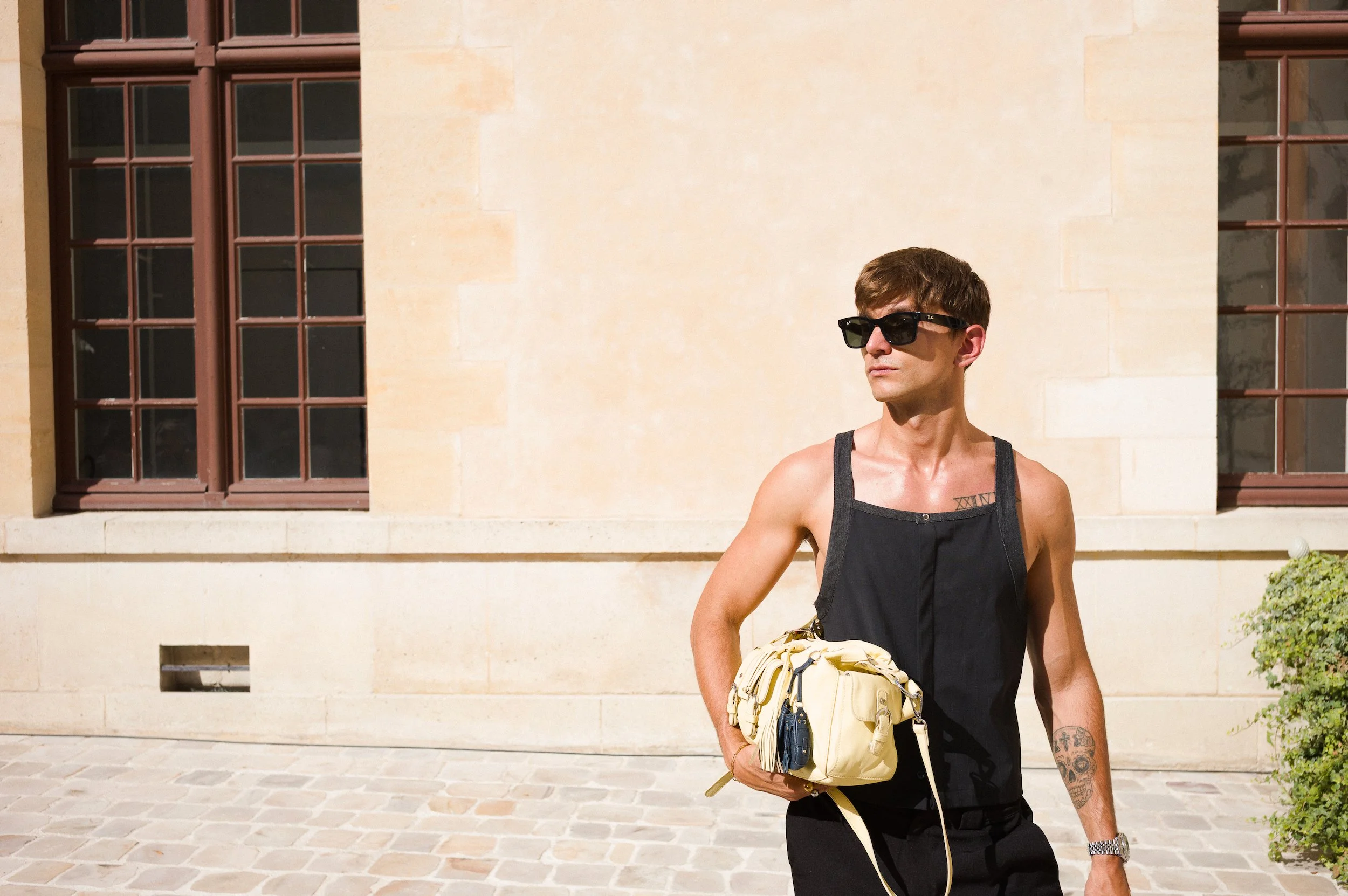 A man with short brown hair, wearing sunglasses, a black sleeveless top, and black pants, stands outside in front of a light-colored building with large windows and a cobblestone ground. He holds a yellow backpack.
