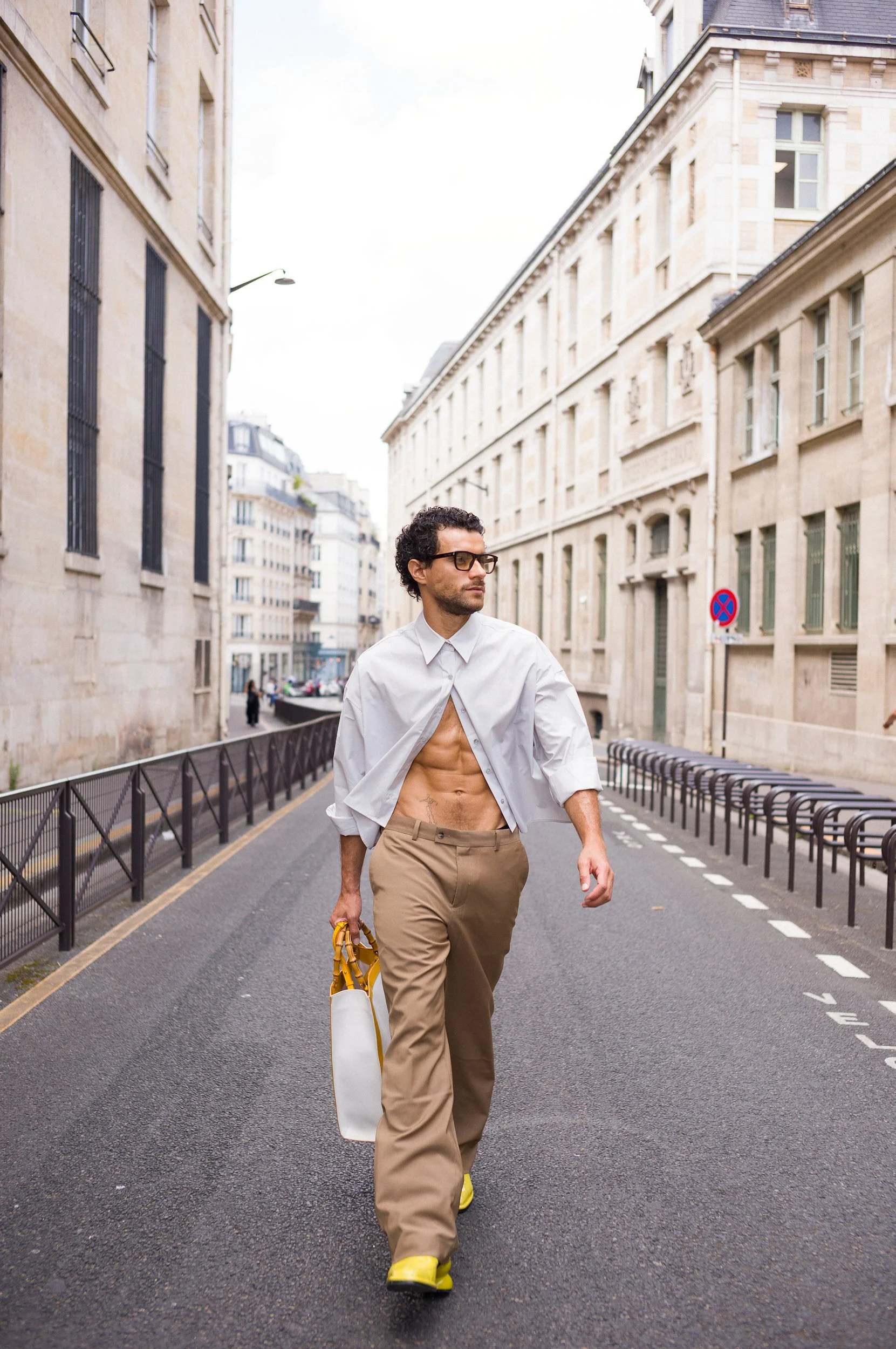 At Paris Fashion Week, A man with curly hair, glasses, and a beard walking on an urban street. He is wearing a white shirt, khaki pants, and yellow shoes, and is carrying a white bag and a yellow item in his right hand.