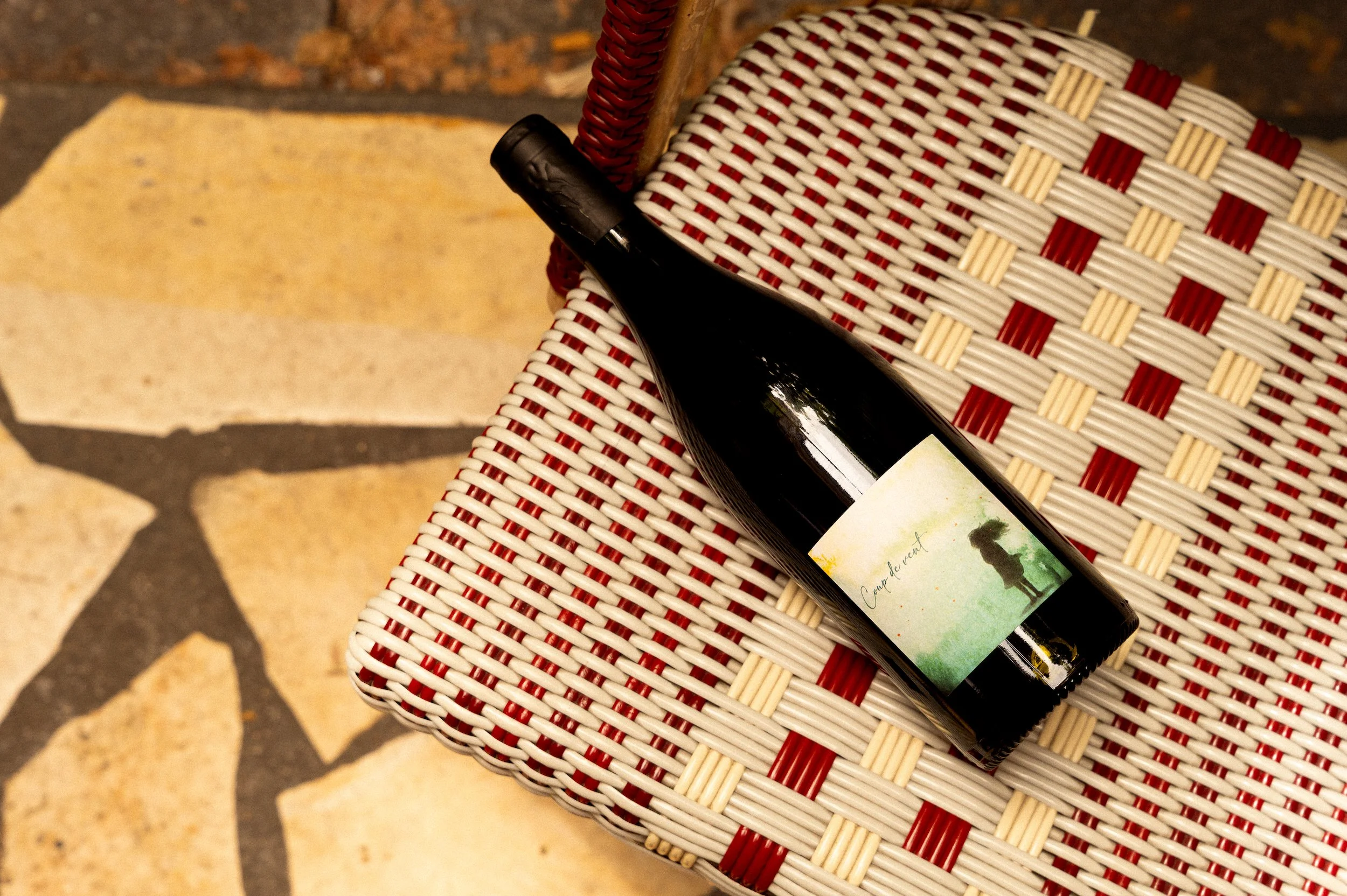 A close-up of a wine bottle resting on a red and white woven chair with a brick floor in the background.