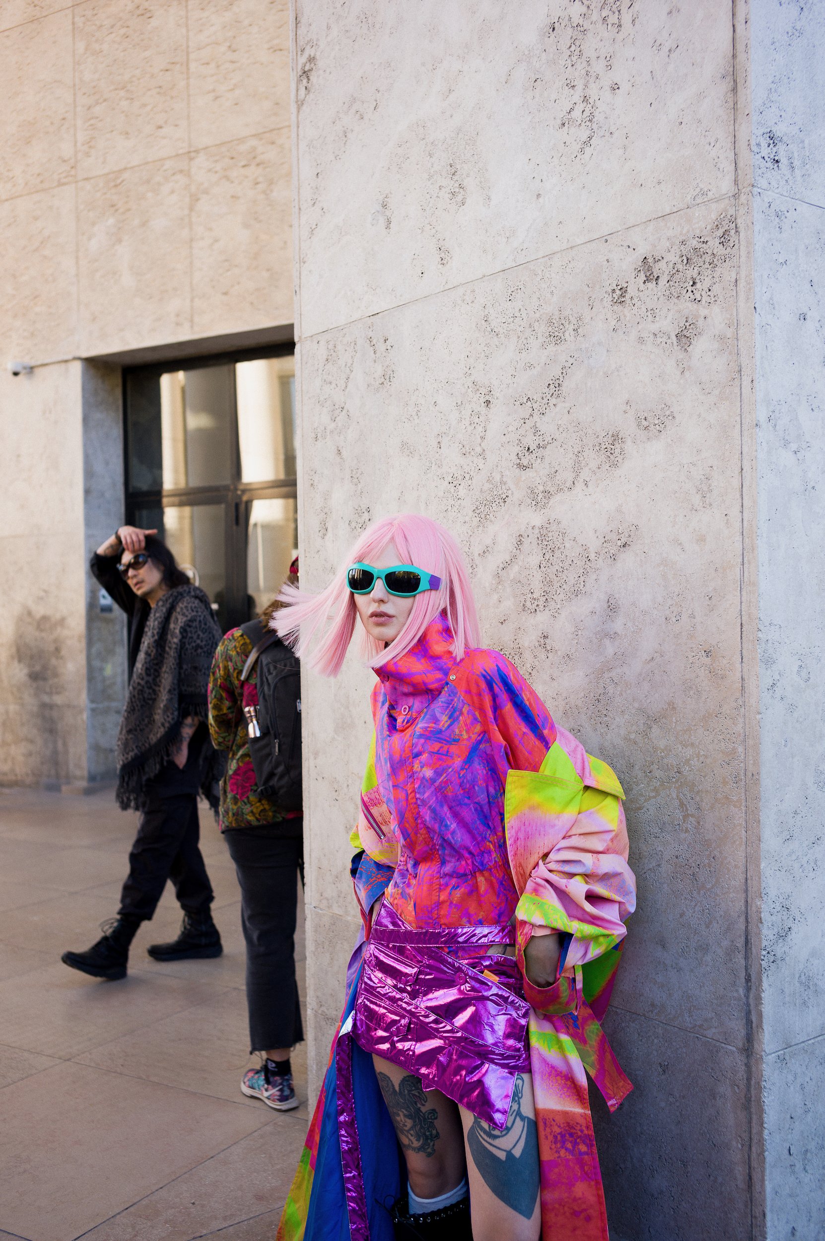 A woman with pink hair, wearing vibrant sunglasses and a colorful jacket, is leaning against a beige stone wall. Two other people are in the background, one with dark hair and glasses, and the other with a patterned coat and dark hair.