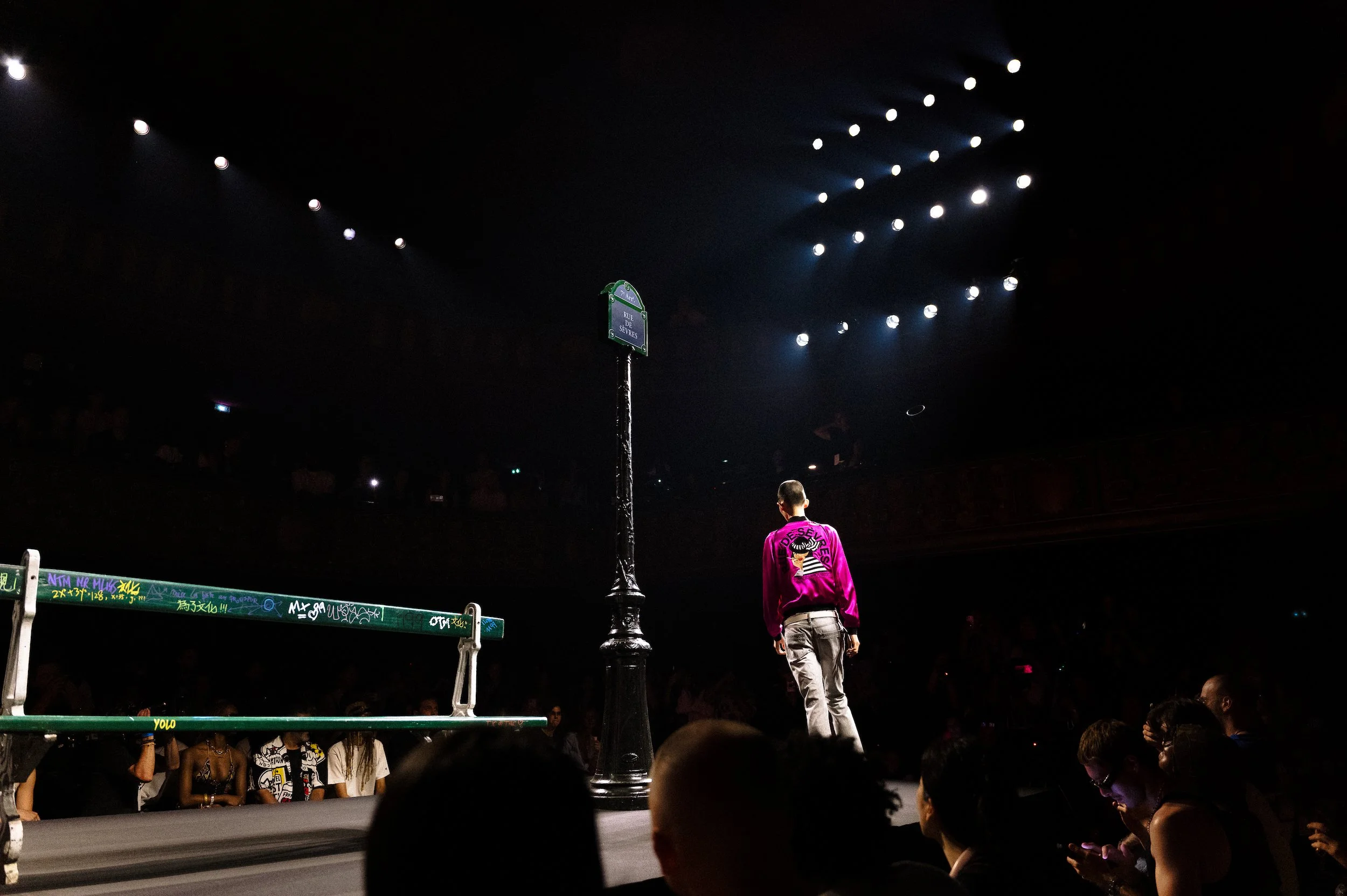 At Paris Fashion week a model during a fashion show, seen from behind, with graffiti-covered barrier and a street sign reading 'Rue des Sevres' in the background, stage lights shining down, and audience members in the dark.