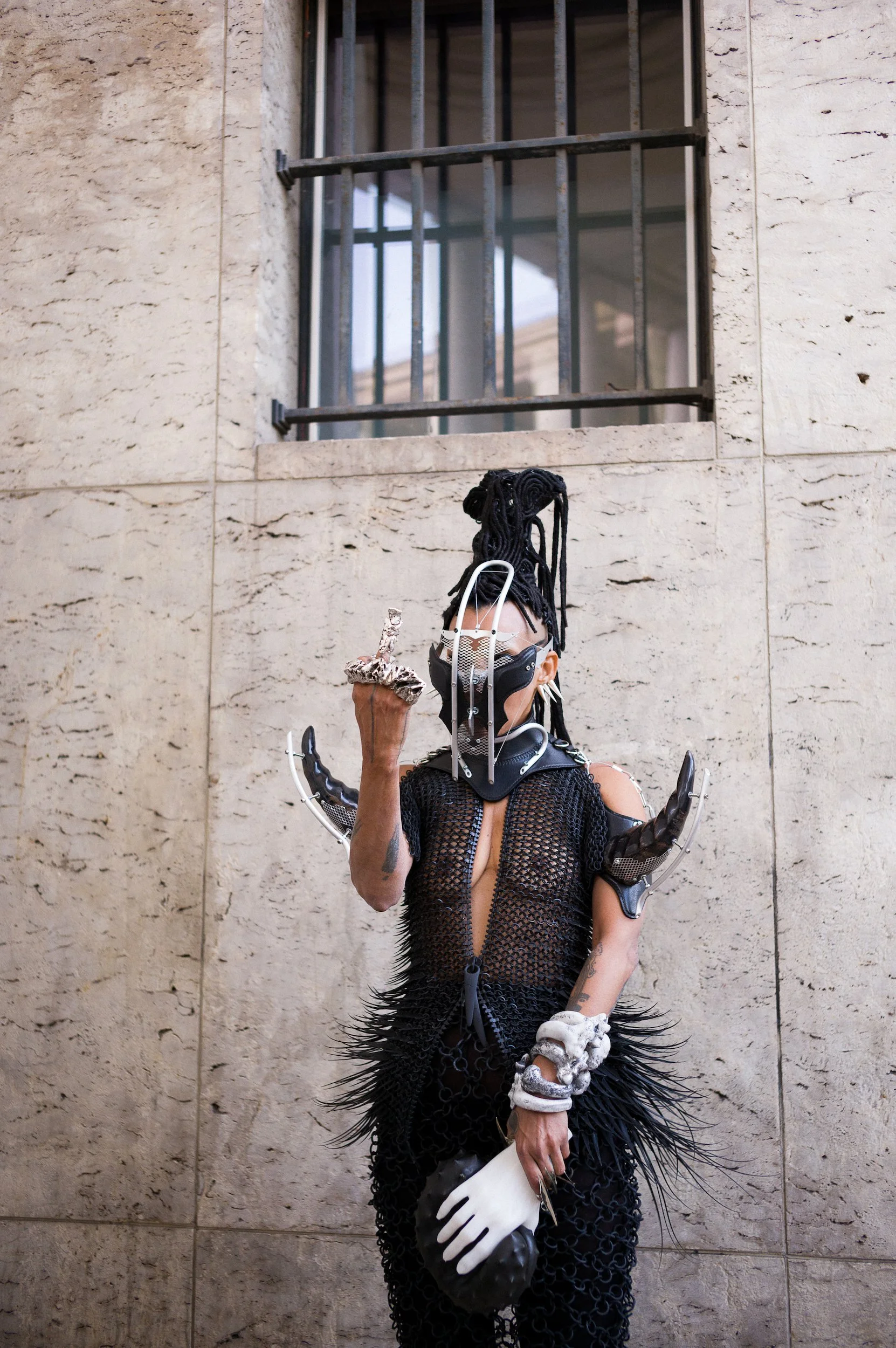 A person with long black dreadlocks wearing a black mesh dress and white gloves, standing against a beige stone wall, holding a skull in one hand and a piece of fabric in the other, with various metallic and robotic accessories on their arms.