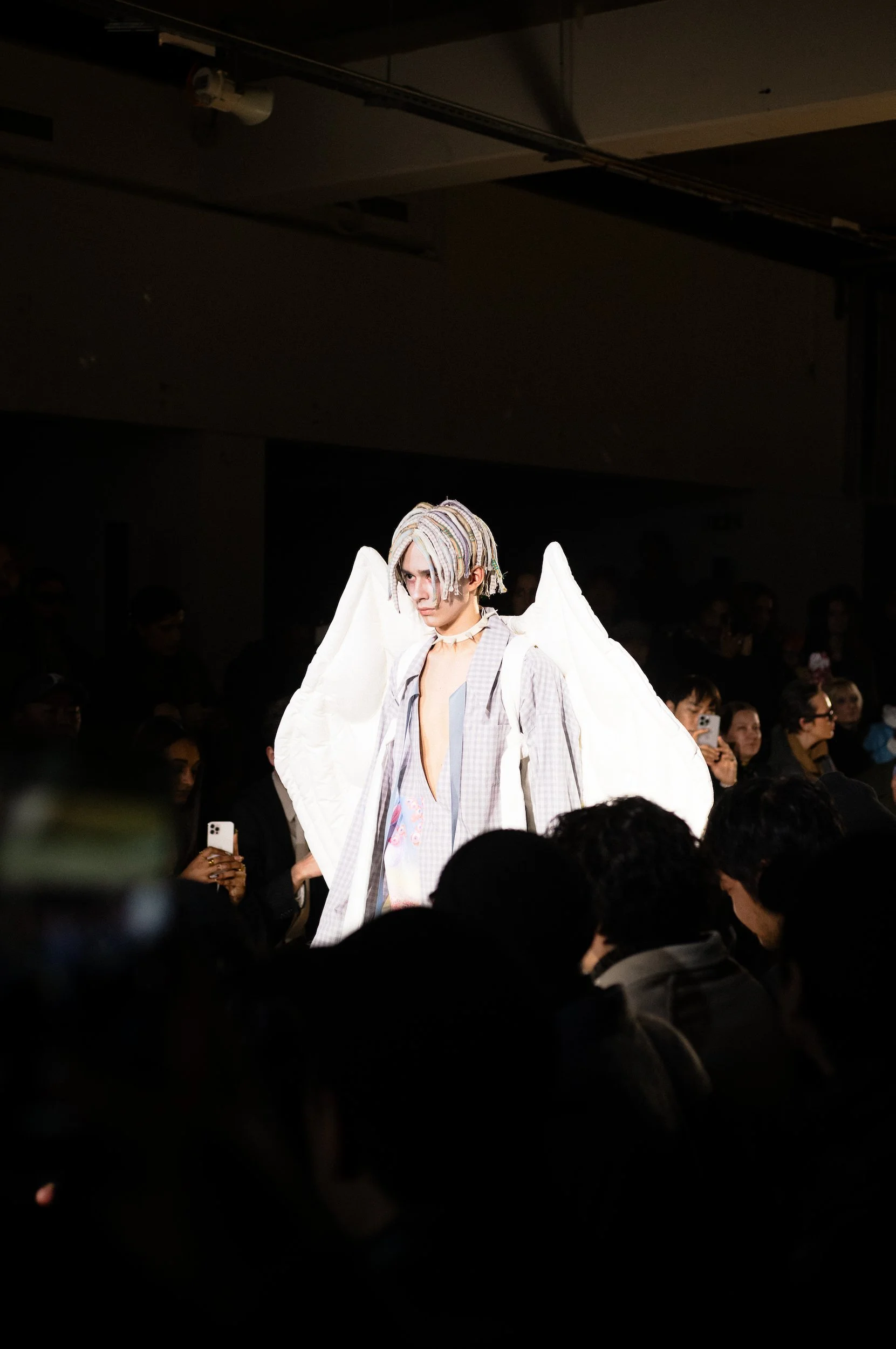 At Paris Fashion Week, A model with pastel-colored dreadlock hair, wearing a plaid jacket with angel wings on his back, walking on a runway during a fashion show with an audience watching.