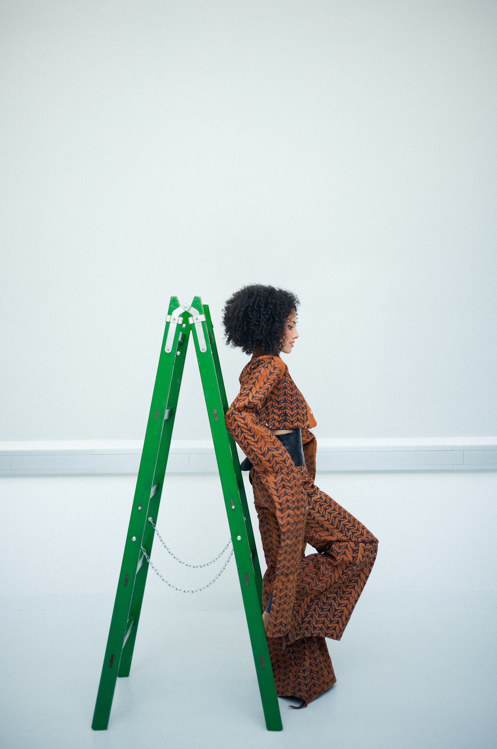 A woman with curly hair wearing a matching brown patterned outfit, standing beside a green ladder against a plain white wall.