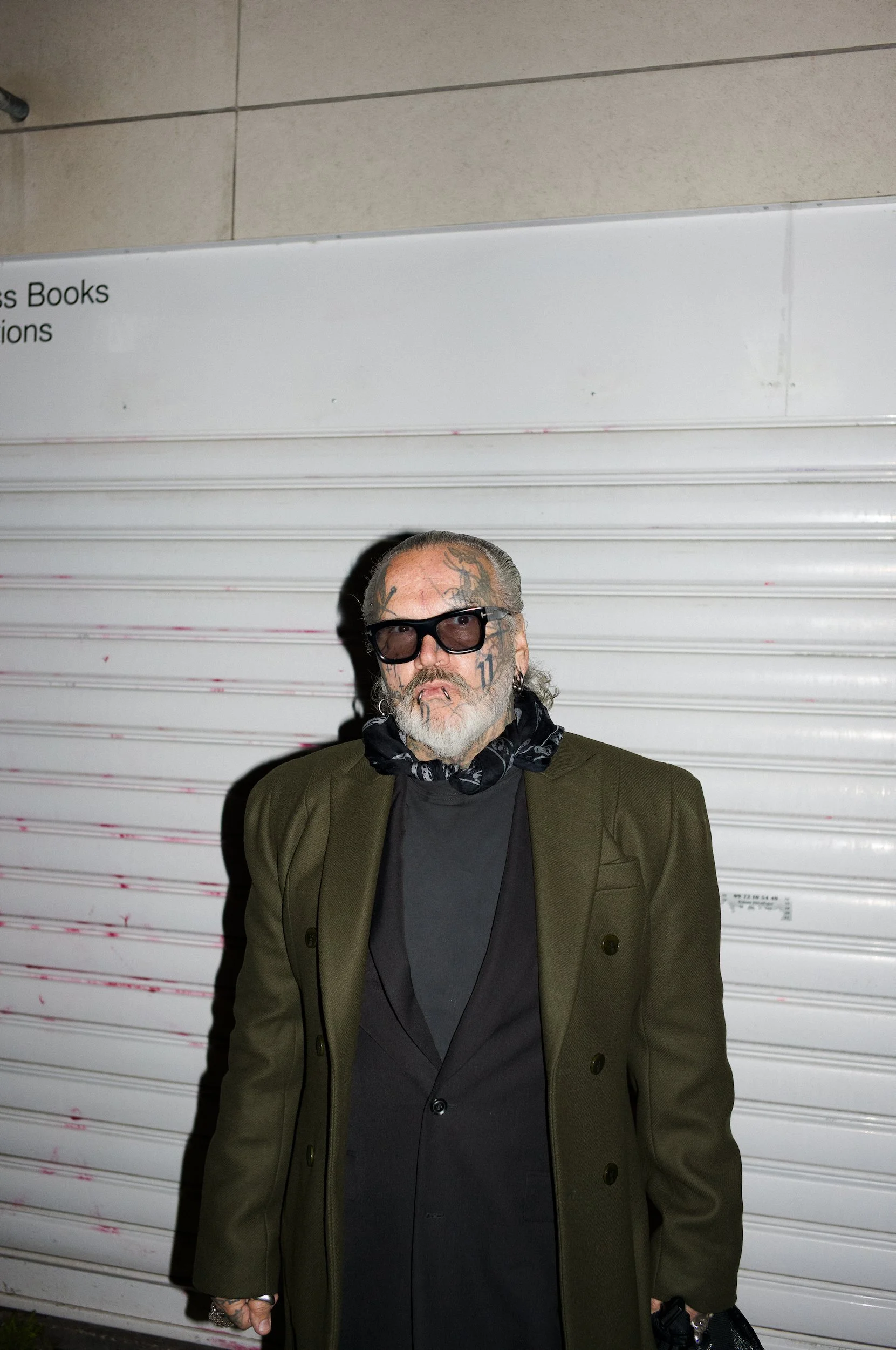 At Paris Fashion Week, Front view of a man with tattoos on his face and hands, wearing dark sunglasses, an olive green blazer, and a black shirt, standing in front of a metal roll-up door at night.